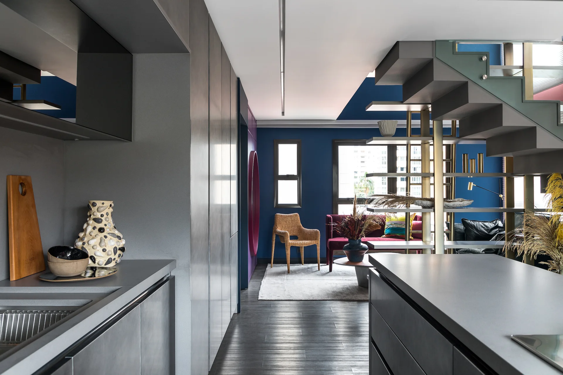 A modern gray kitchen leads to a blue living room featuring a magenta sofa, a wicker chair, and a metal staircase with integrated shelving.