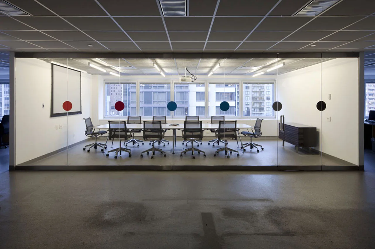 A glass-walled office conference room features a long white table surrounded by ergonomic rolling chairs under a grid ceiling.