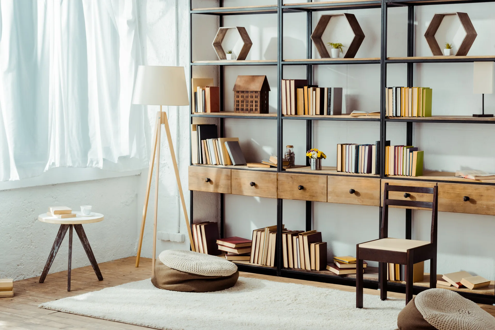 A large black metal and wood bookshelf stands next to a wooden tripod lamp and side table on a light area rug.