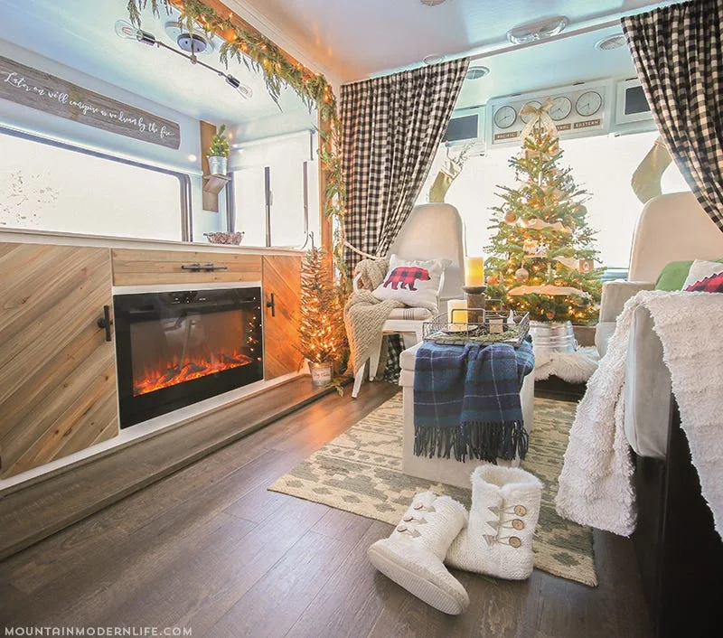 A cozy RV interior decorated for Christmas with a built-in electric fireplace, herringbone wood cabinetry, and buffalo plaid curtains.