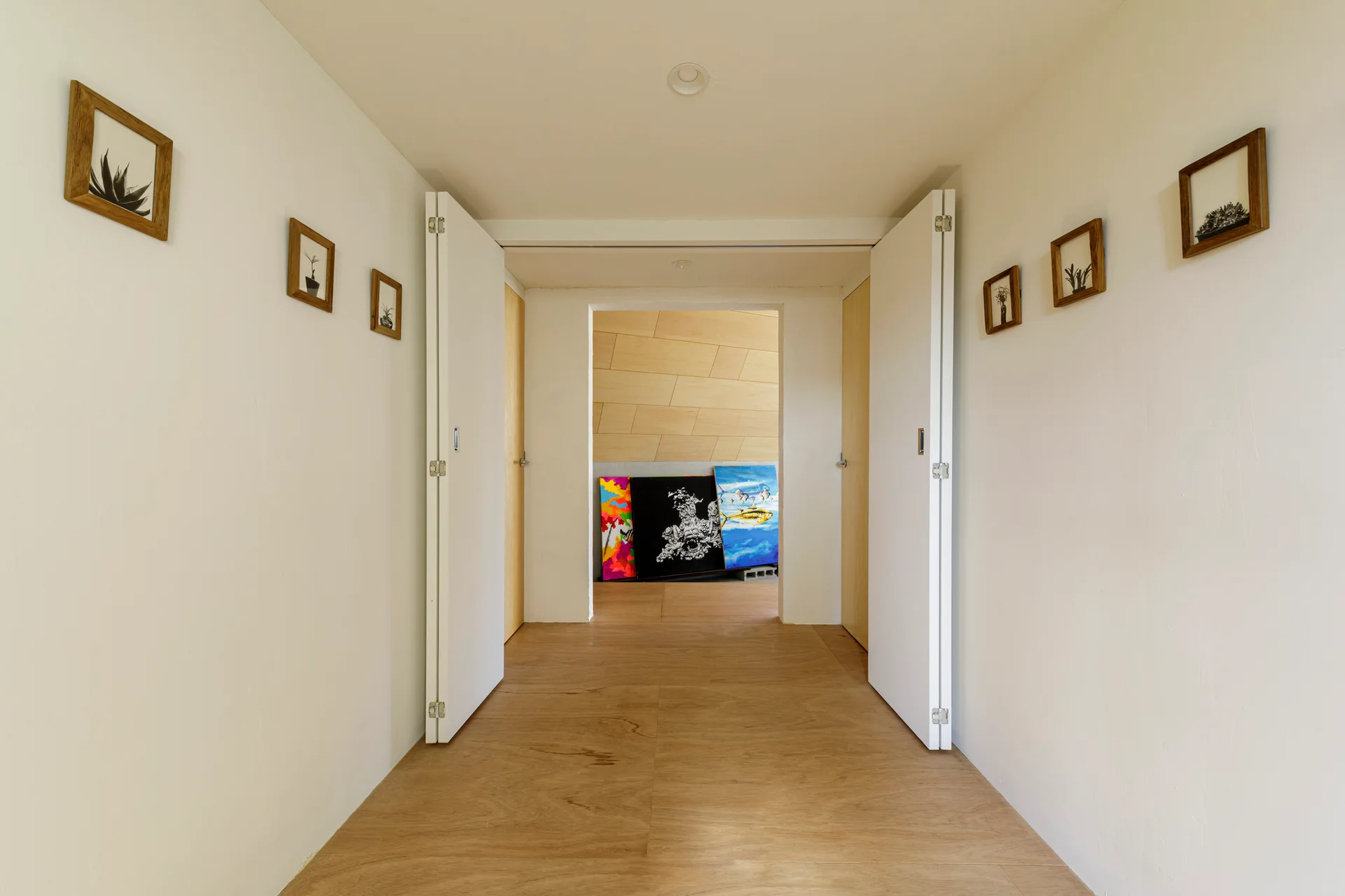 A symmetrical white hallway with light wood floors features small wood-framed pictures on the walls and open white bifold doors.