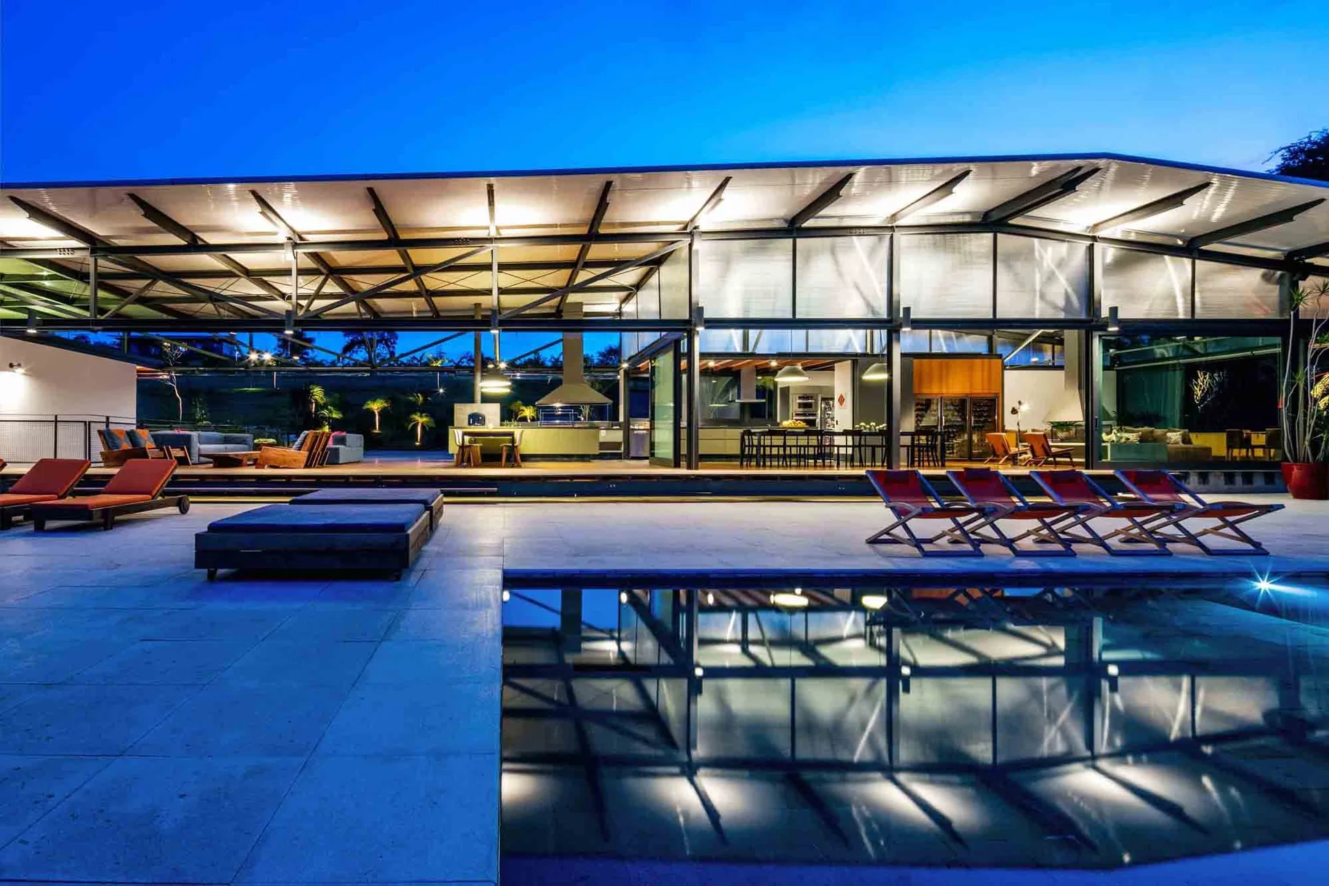 A modern open-air pavilion with steel beams features a lounge and dining area next to a swimming pool with lounge chairs at twilight.