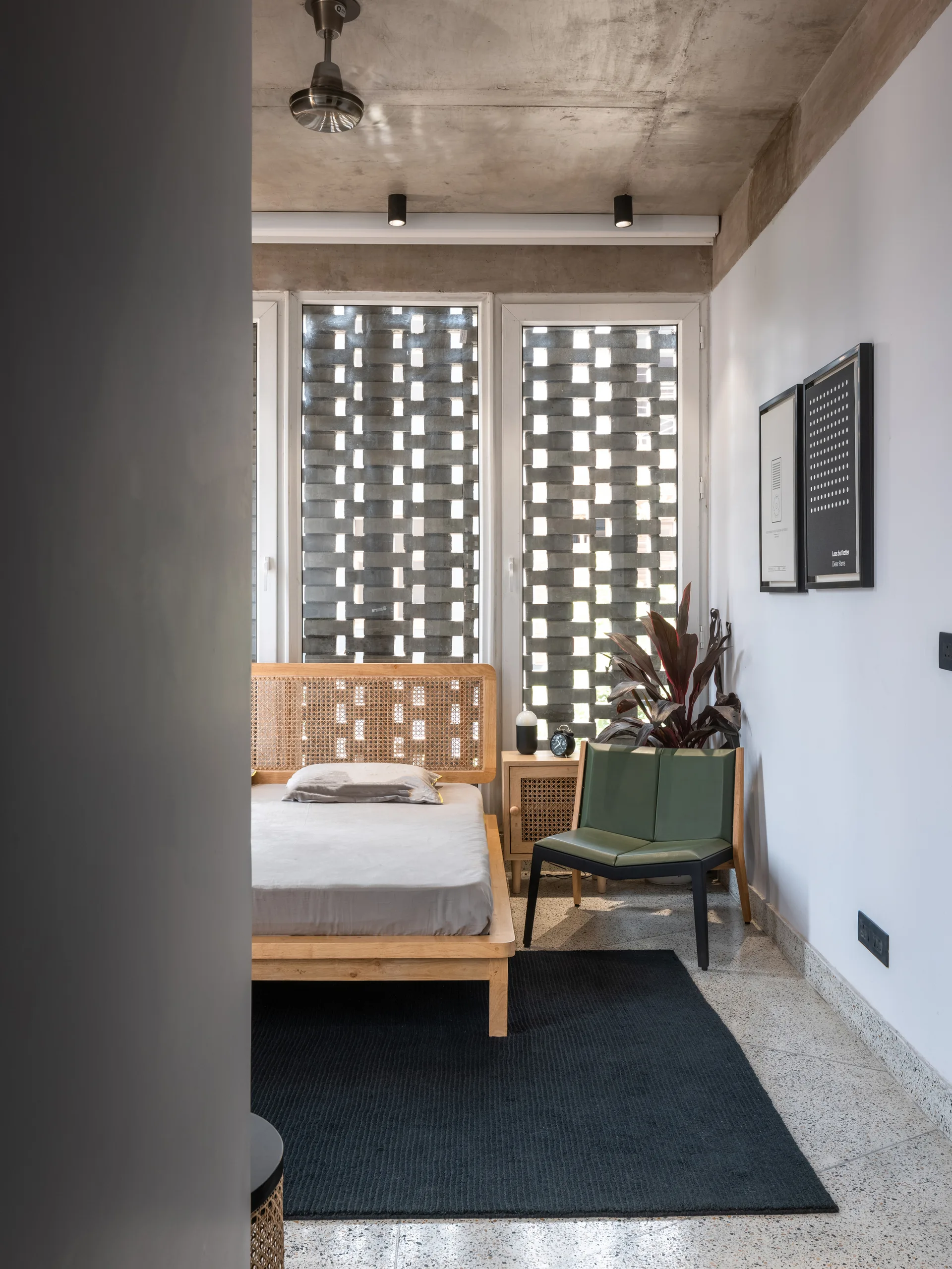A modern bedroom features a light wood bed with a woven rattan headboard, a dark grey rug on a terrazzo floor, and an exposed concrete ceiling.