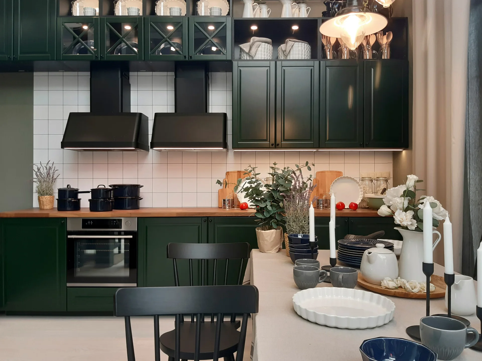 Dark green kitchen cabinets are paired with a white square-tile backsplash, wood countertops, and a dining table set with white and blue dishware.