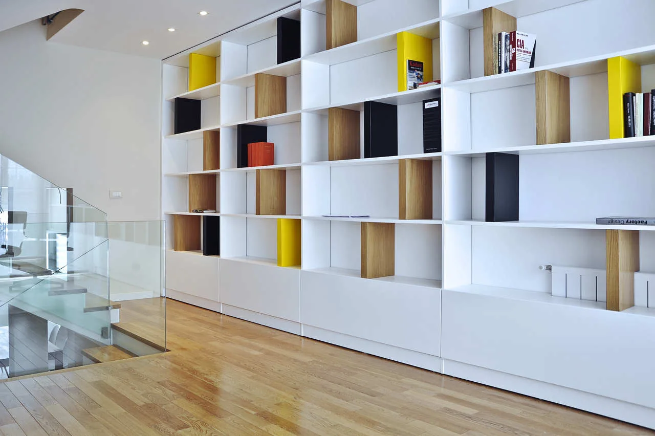 A large white floor-to-ceiling modular bookshelf with wood-toned and yellow inserts stands next to a wooden staircase with a clear glass railing.