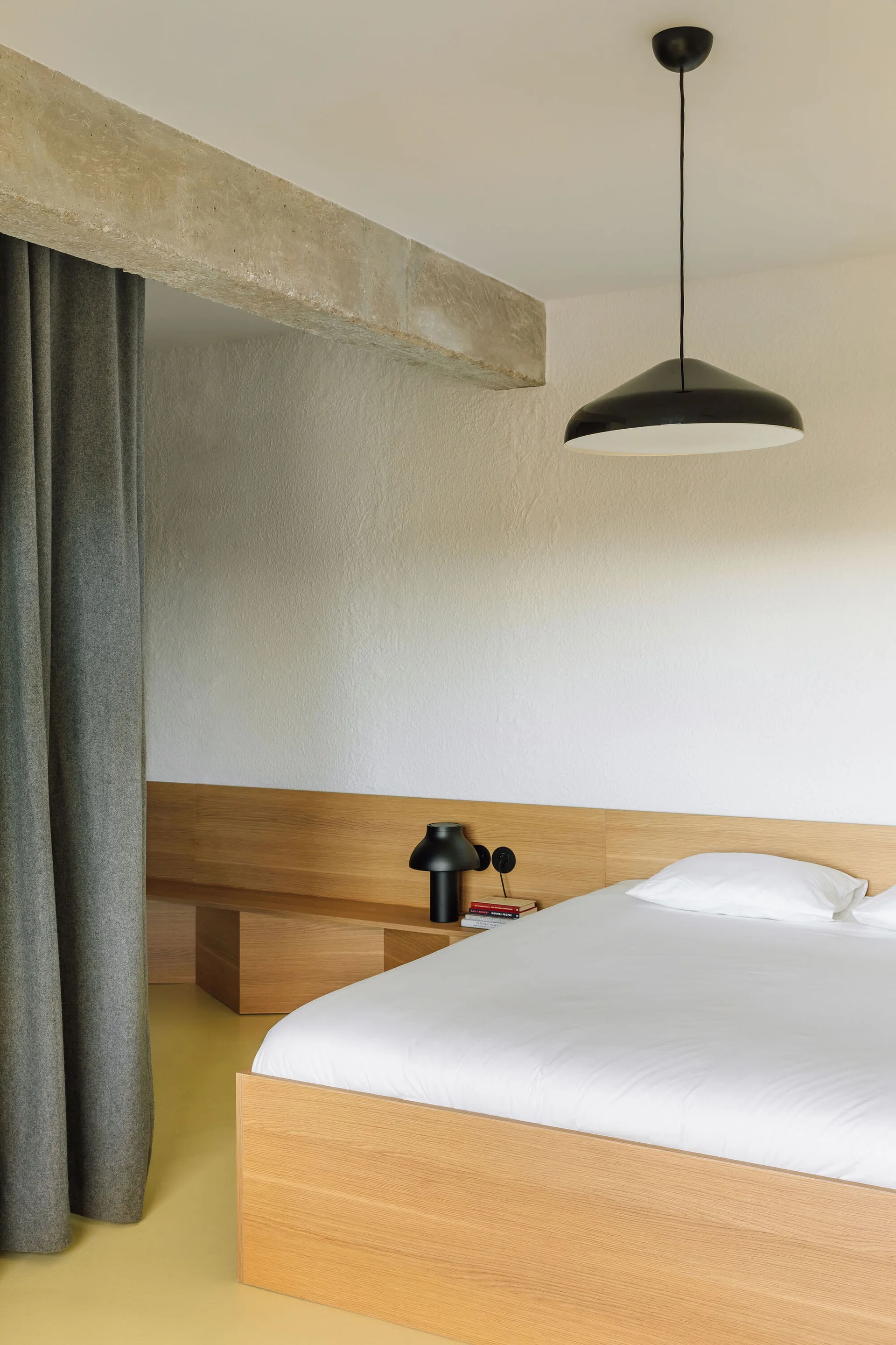 A minimalist bedroom features a wooden platform bed with an extended headboard, a black pendant light, a grey curtain, and an exposed concrete beam.