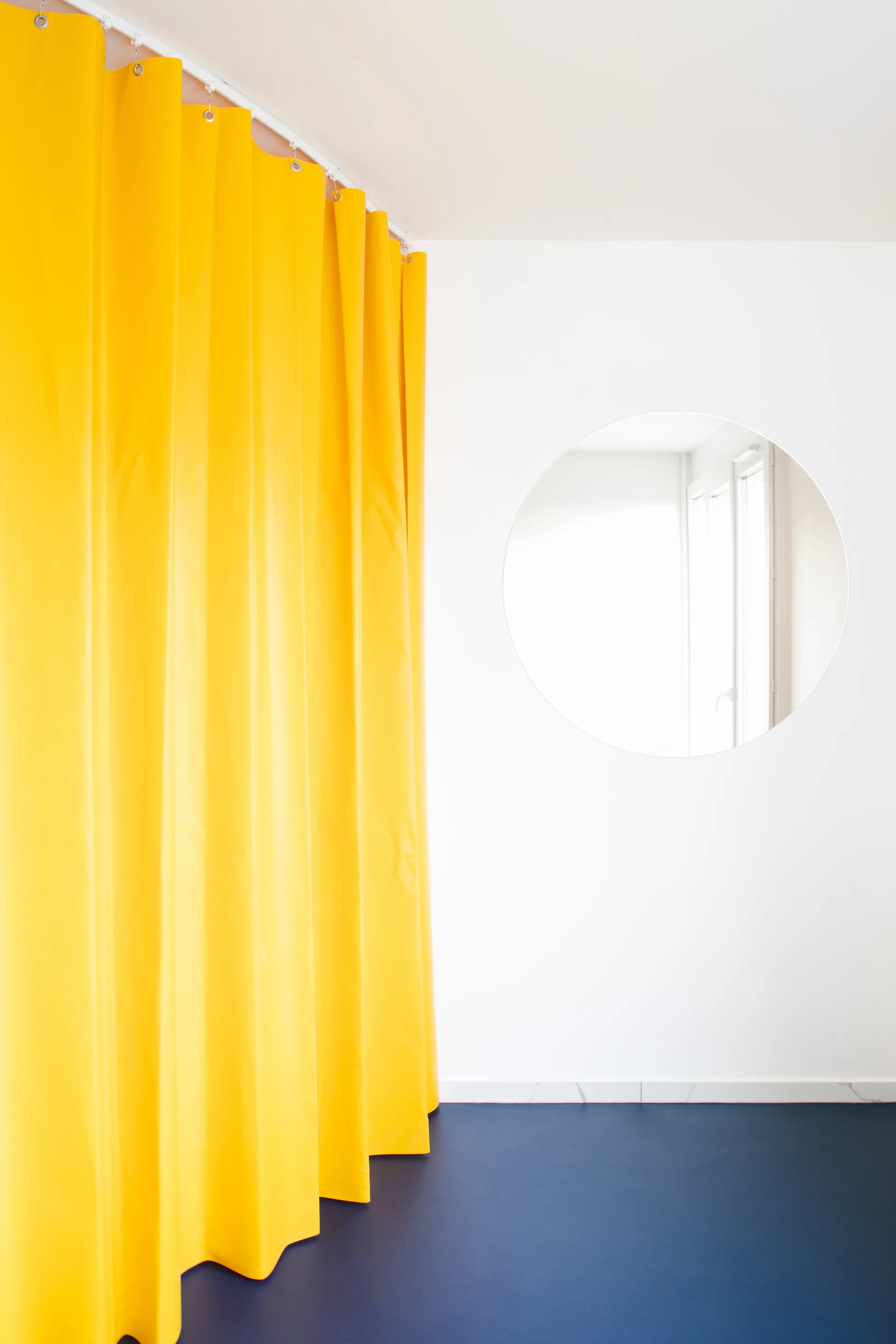 A bright yellow floor-length curtain hangs on a ceiling track next to a large circular frameless mirror on a white wall with a blue floor.