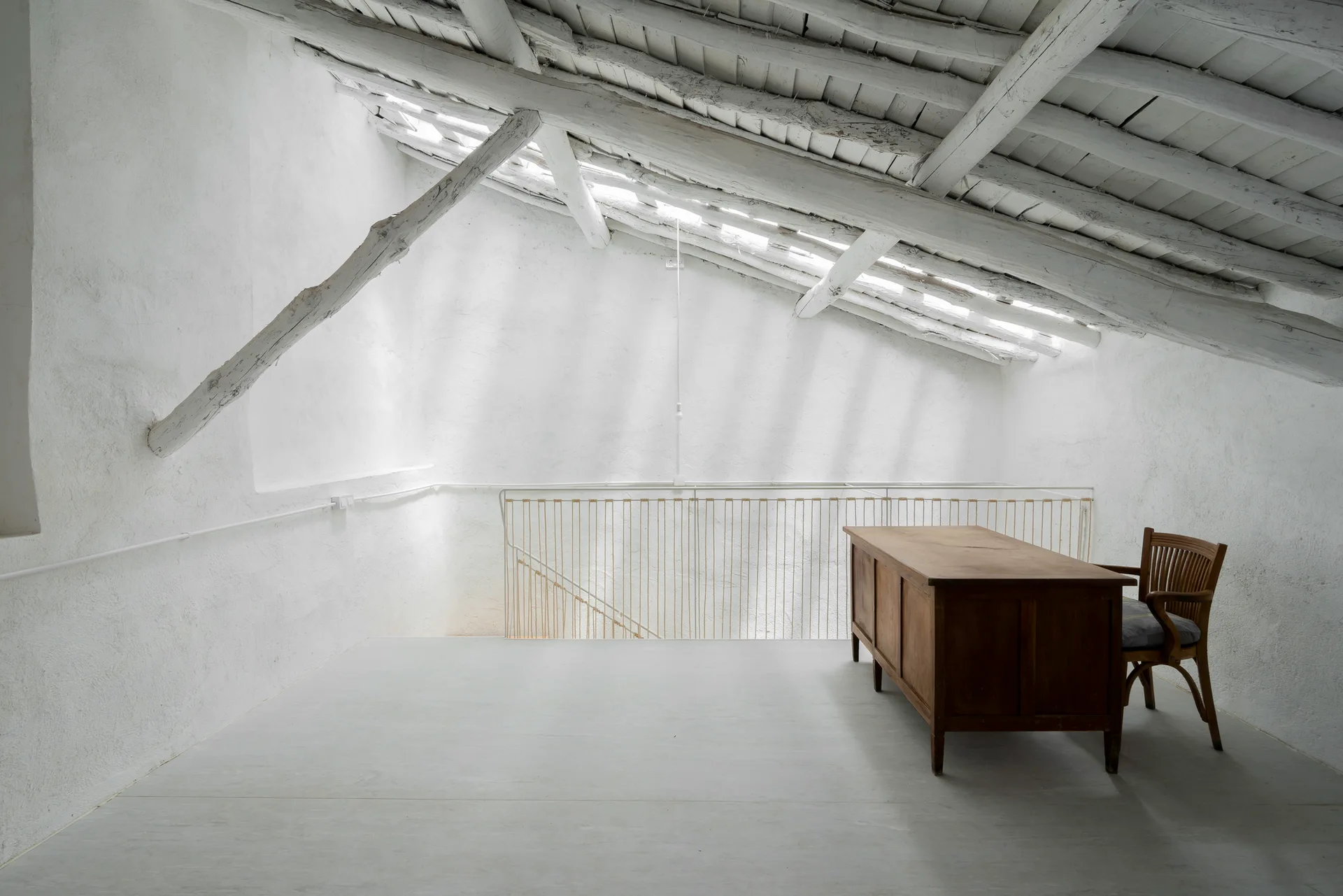 A rustic wooden desk and chair sit on a light grey floor beneath white textured plaster walls and a sloped ceiling with painted timber beams.
