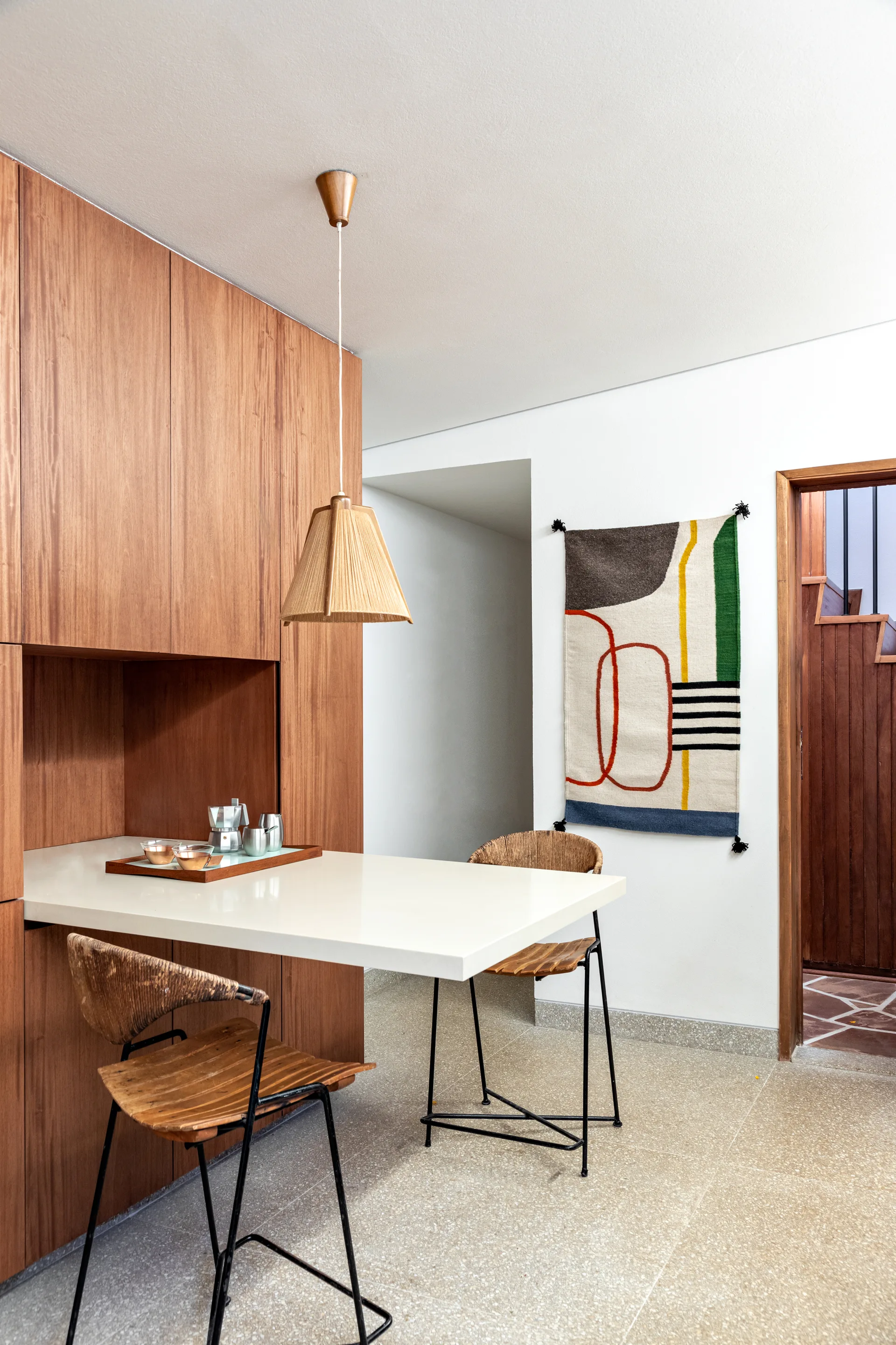 A white breakfast bar extends from built-in wood cabinets, featuring two wooden stools, a woven pendant light, and a colorful geometric wall hanging.