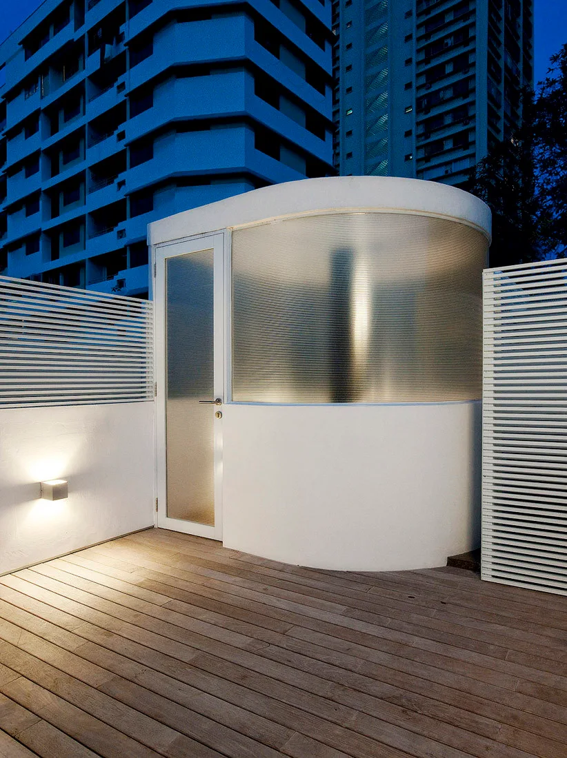 A modern rooftop terrace at night features a curved white structure with ribbed glass panels, wood plank decking, and a slatted privacy screen.