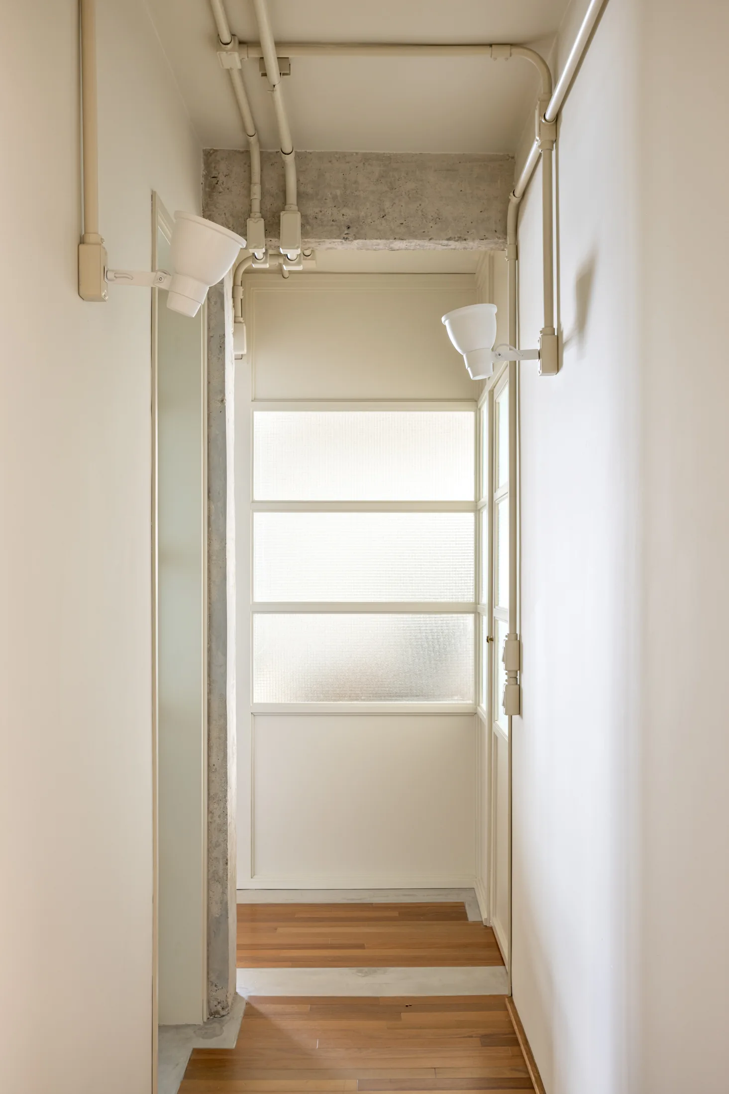 A narrow off-white hallway features wood flooring, exposed painted conduits, a raw concrete beam, and a multi-pane frosted glass door.