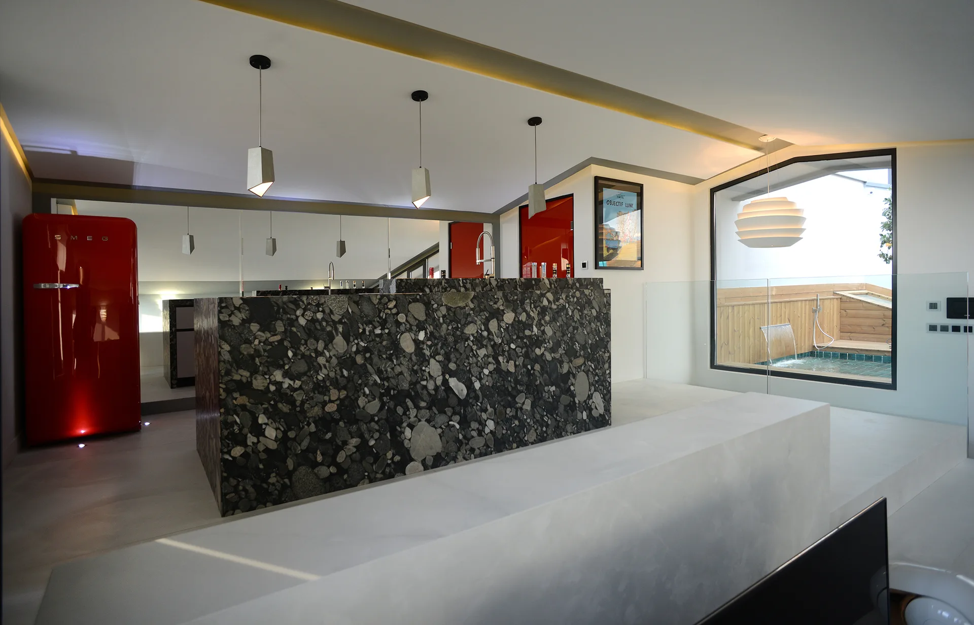 A modern white kitchen featuring a large dark aggregate stone island, concrete pendant lights, and a vibrant red refrigerator.