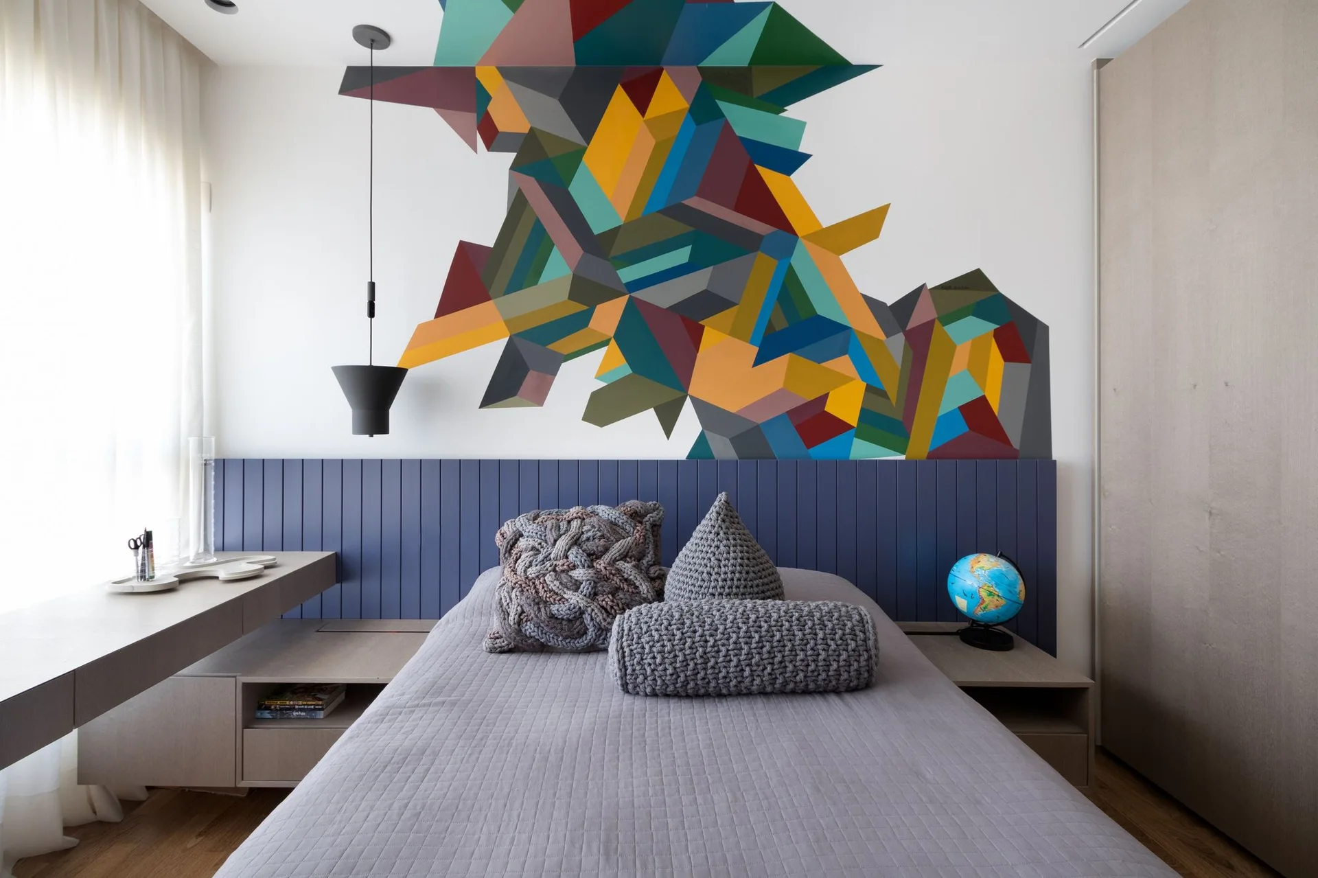 A modern bedroom featuring a gray bed with chunky knit pillows, a blue paneled headboard, and a multicolored geometric mural on the wall.