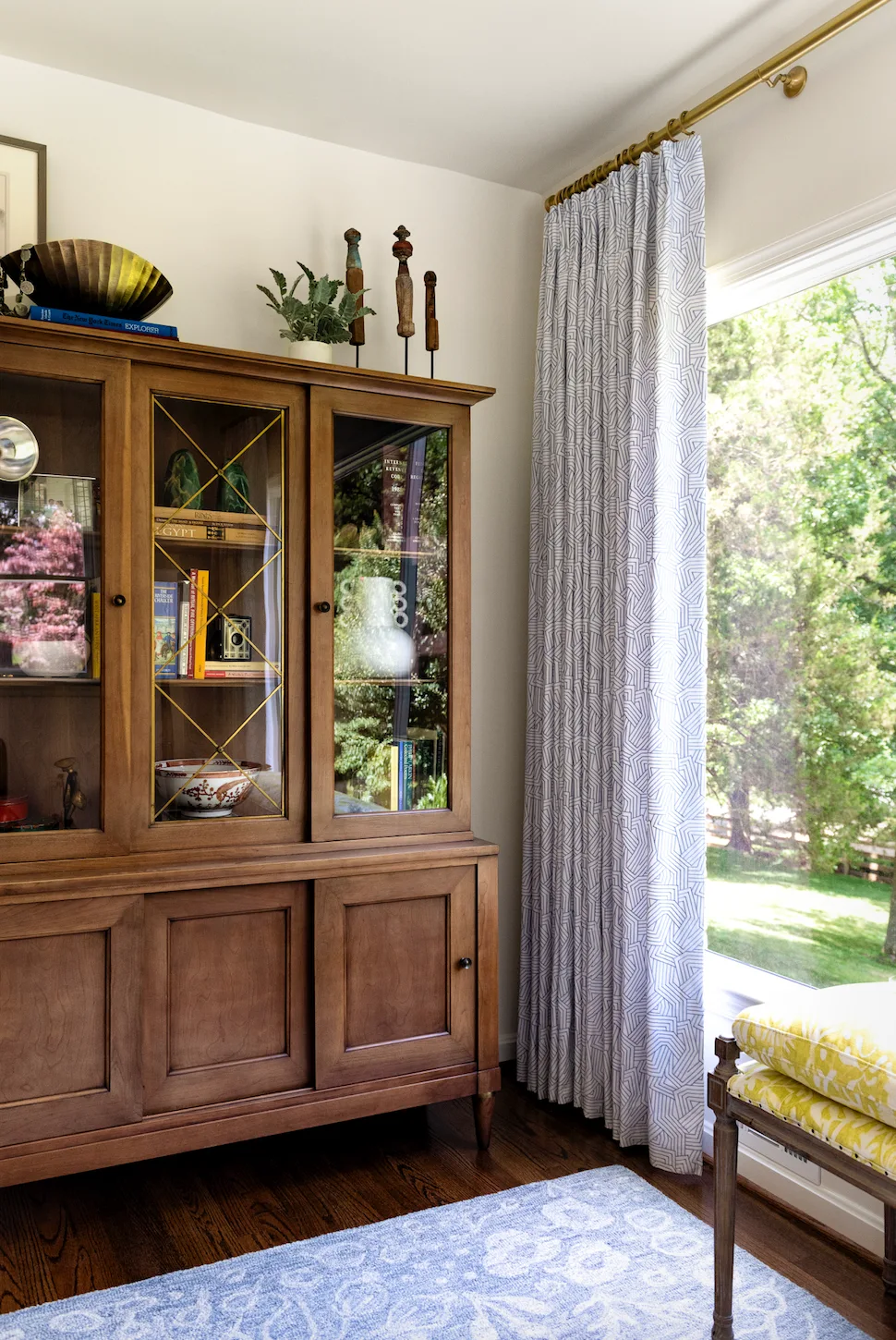 A traditional wood cabinet filled with books and pottery sits beside a tall window dressed in blue and white patterned curtains.