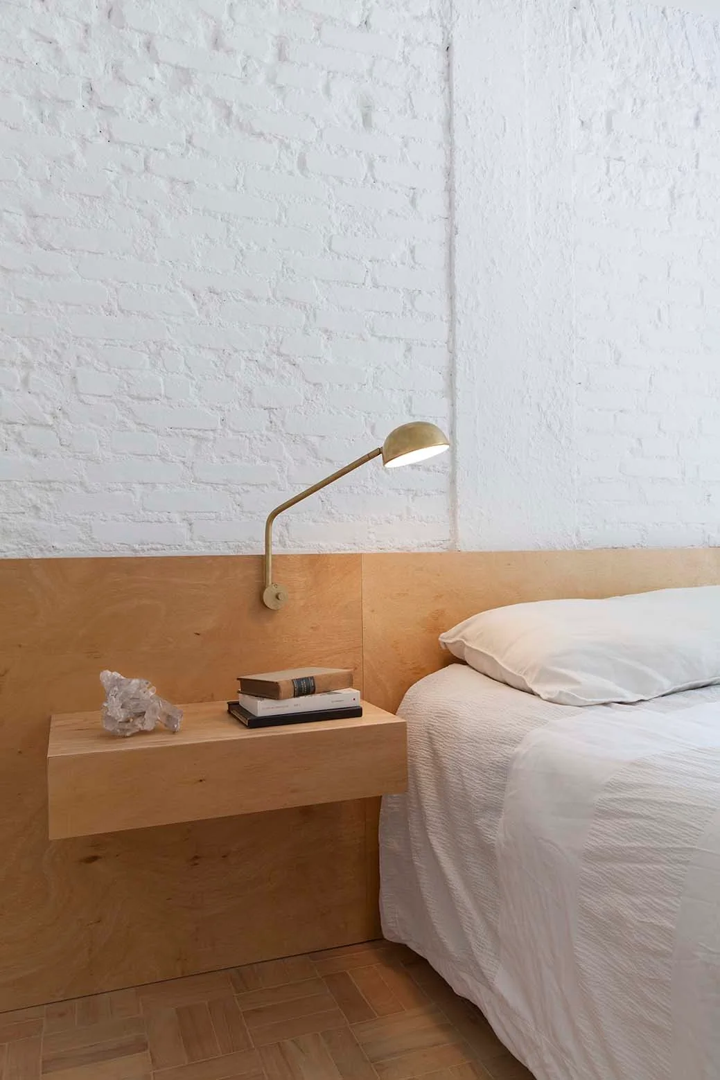 A minimalist bedroom features a bed with white linens, a floating wood nightstand, a brass sconce, and a wall of white brick and plywood paneling.