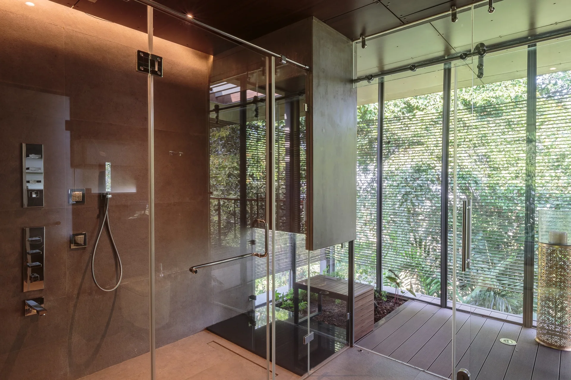 A modern bathroom features a glass-enclosed shower stall with chrome fixtures next to floor-to-ceiling windows showing lush green plants.
