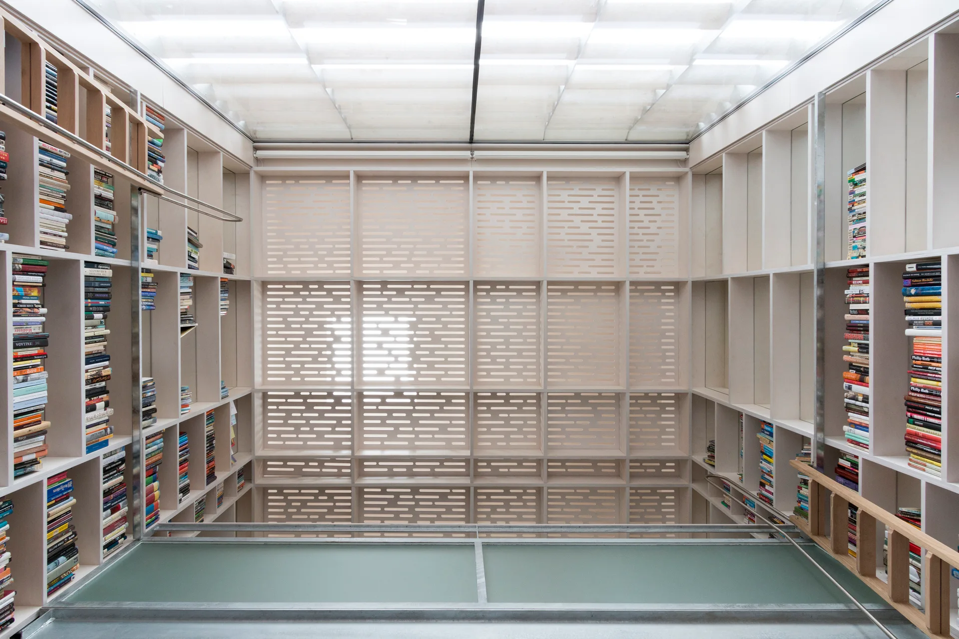A floor-to-ceiling grid of light wood library shelving filled with books under a bright, translucent paneled skylight.