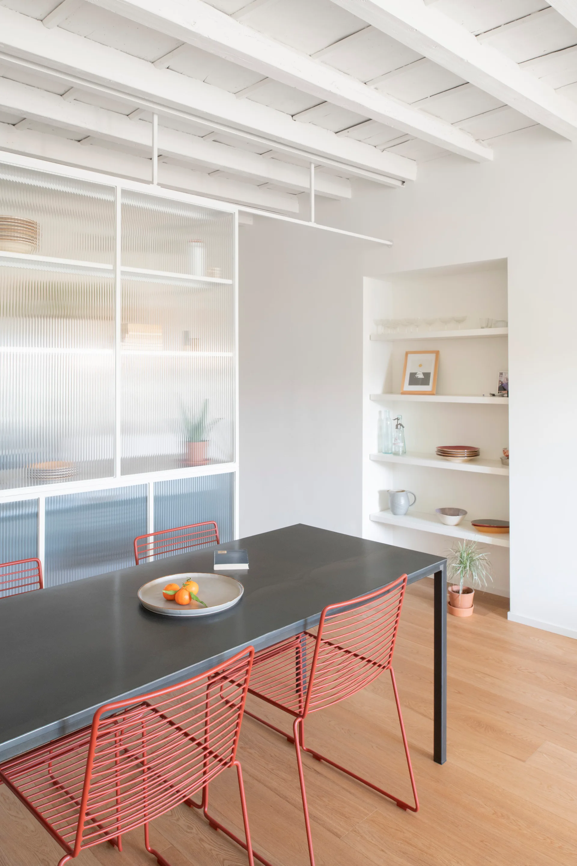 A black dining table with red wire chairs sits in a bright white room featuring a fluted glass partition and built-in wall shelves.