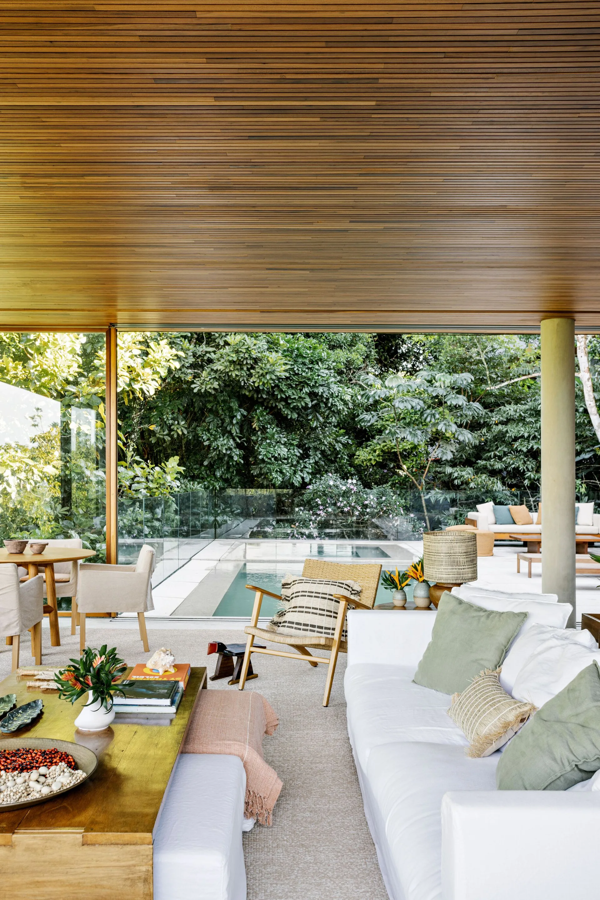 A covered outdoor lounge features a white sofa, low wooden coffee table, and wood slat ceiling looking out toward a swimming pool.