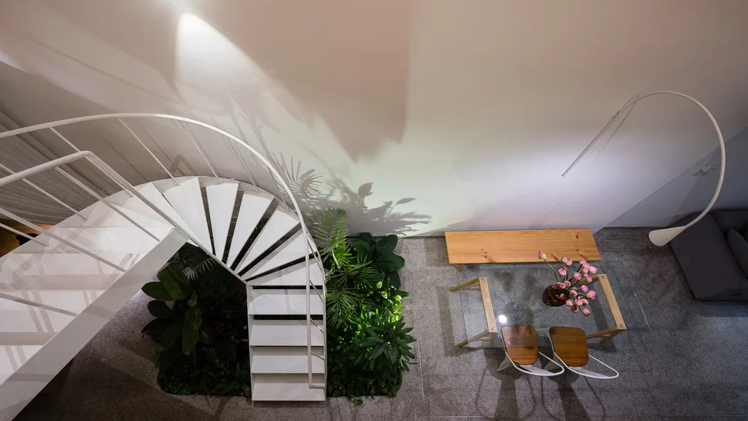 An overhead view of a white spiral staircase curving over an indoor garden bed next to a glass dining table on grey tile flooring.