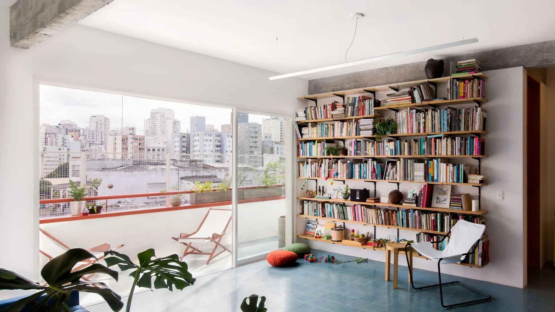 A bright living room features blue tile flooring, a large wall-mounted wooden bookshelf, and sliding glass doors leading to an urban balcony.
