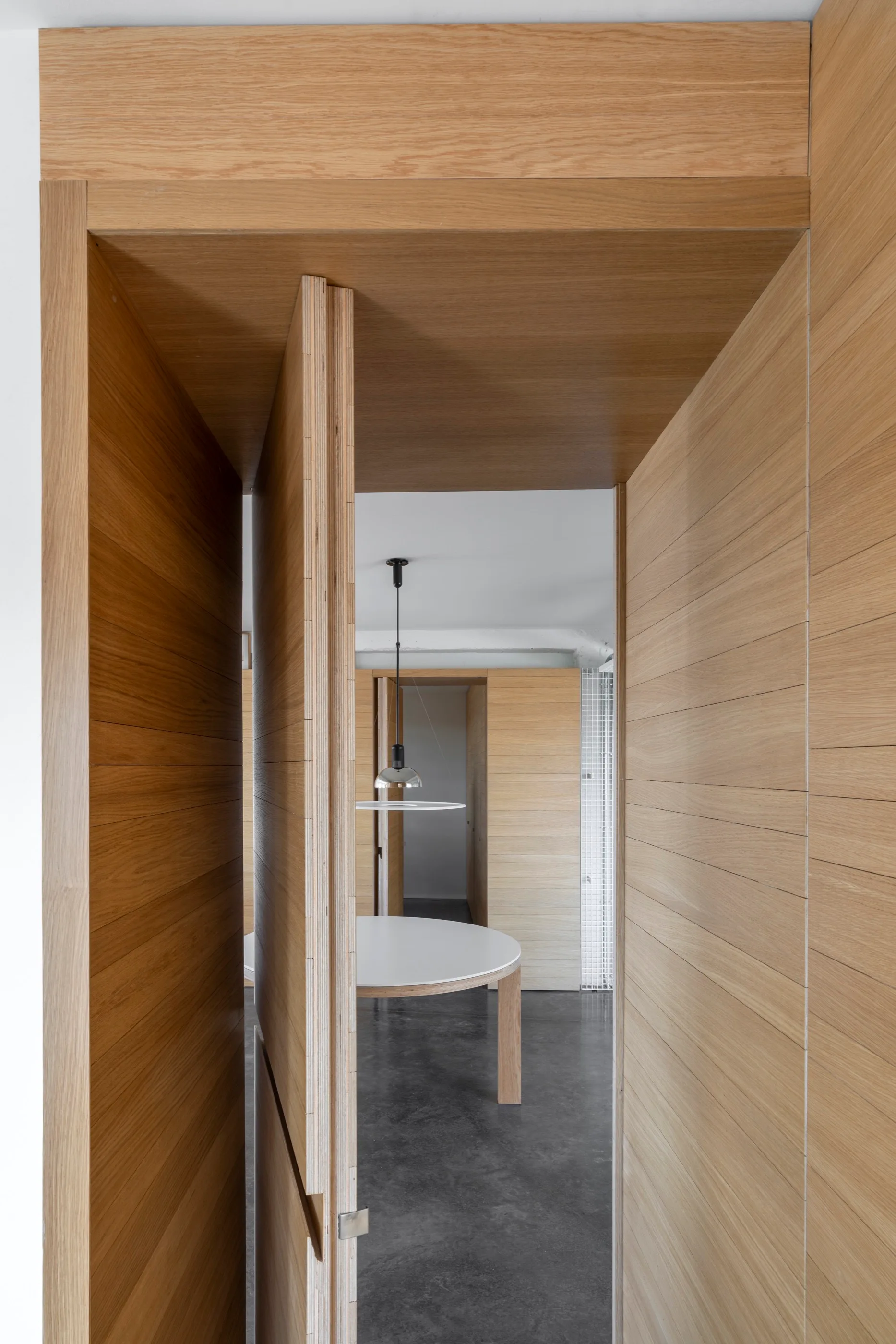 A wood-paneled hallway with an open timber pivot door leads to a minimalist dining room featuring a round white table and dark concrete floors.