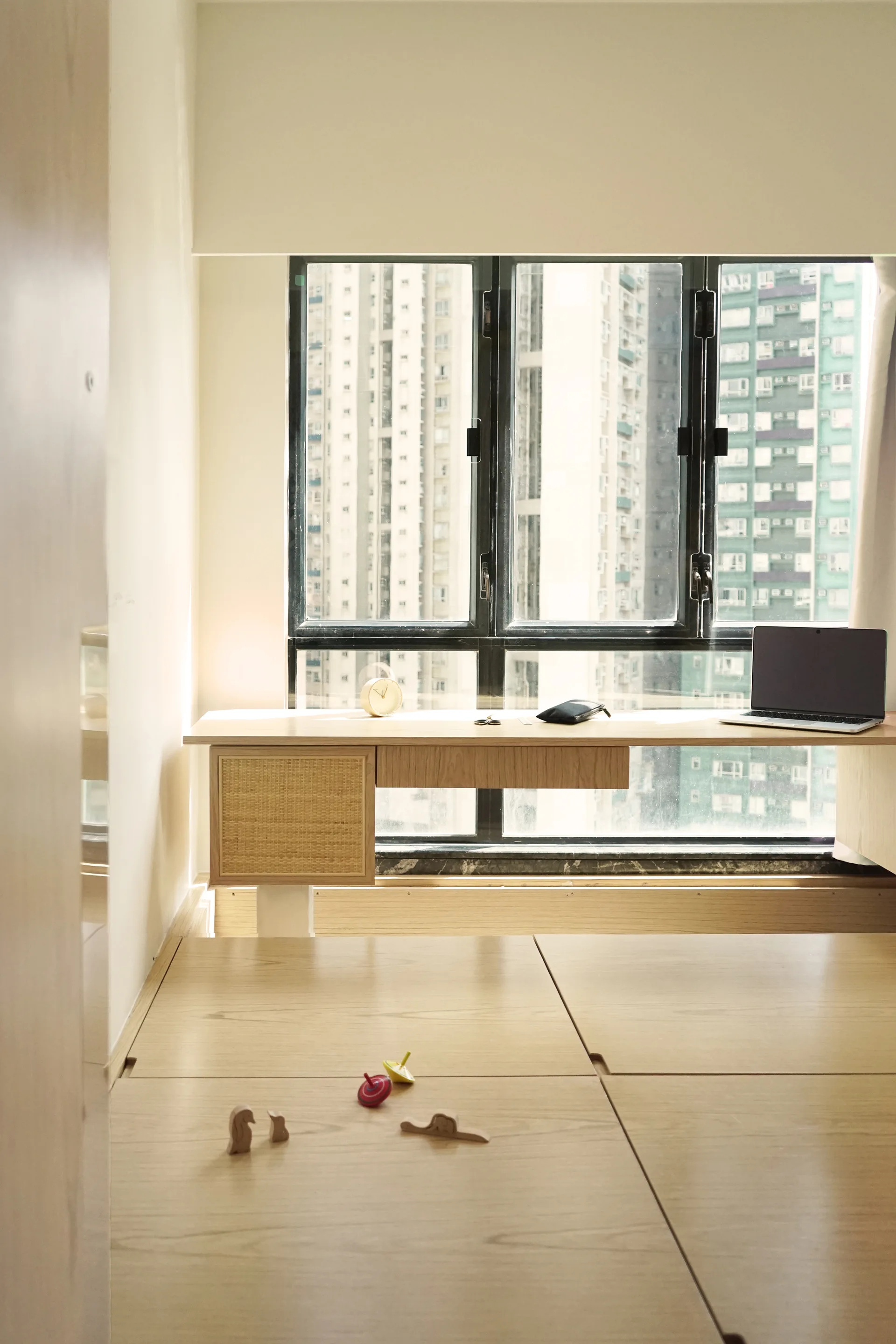 A light wood built-in desk with a rattan drawer front sits before a black-framed window, positioned over a raised wooden platform floor.