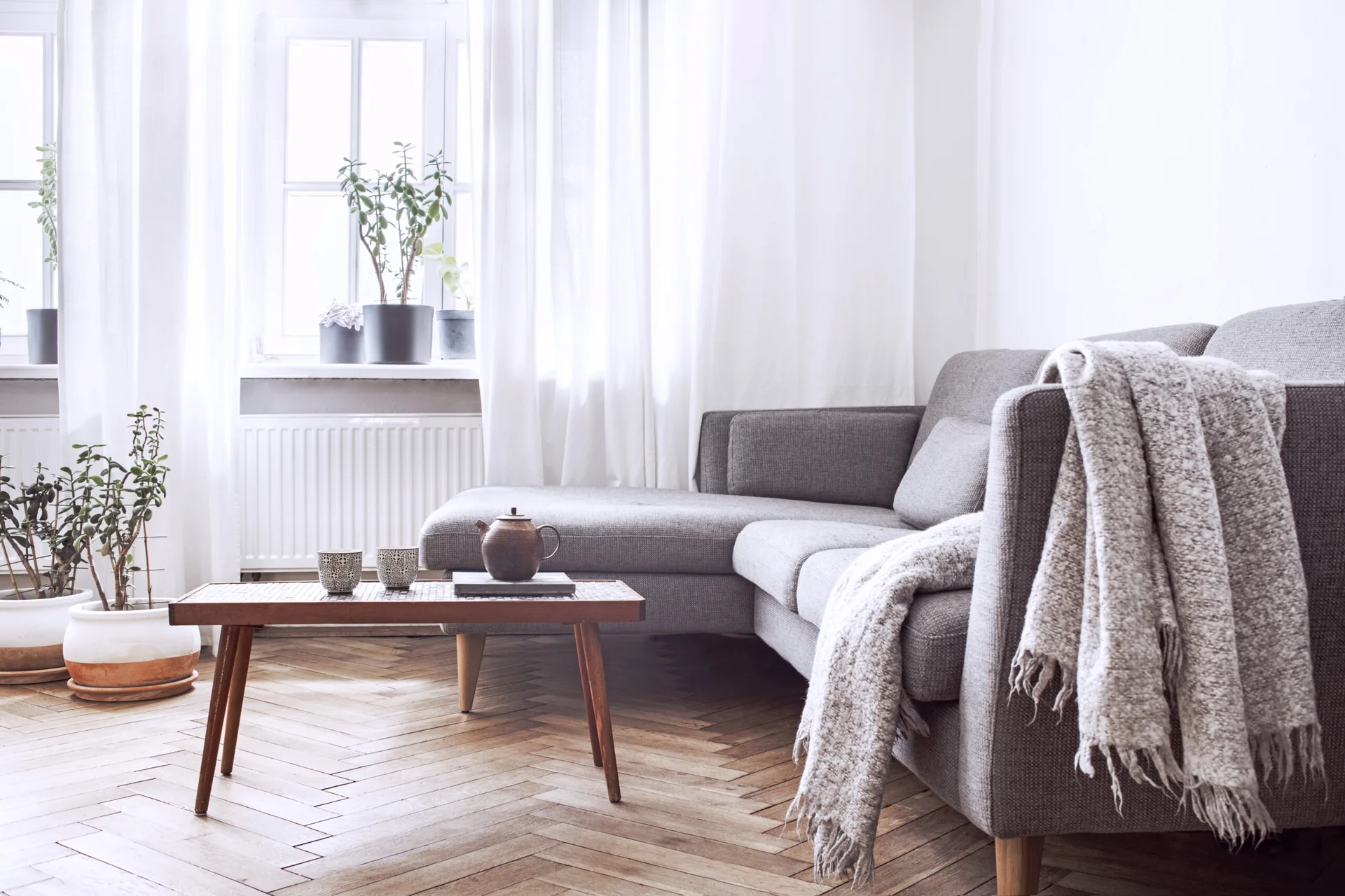 A light-filled living room featuring a grey fabric sectional sofa, a low wooden coffee table, and herringbone parquet wood flooring.