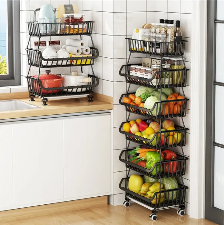 Two black tiered metal wire storage racks stand on a wood countertop and floor filled with fresh produce and jars against a white tiled kitchen wall.
