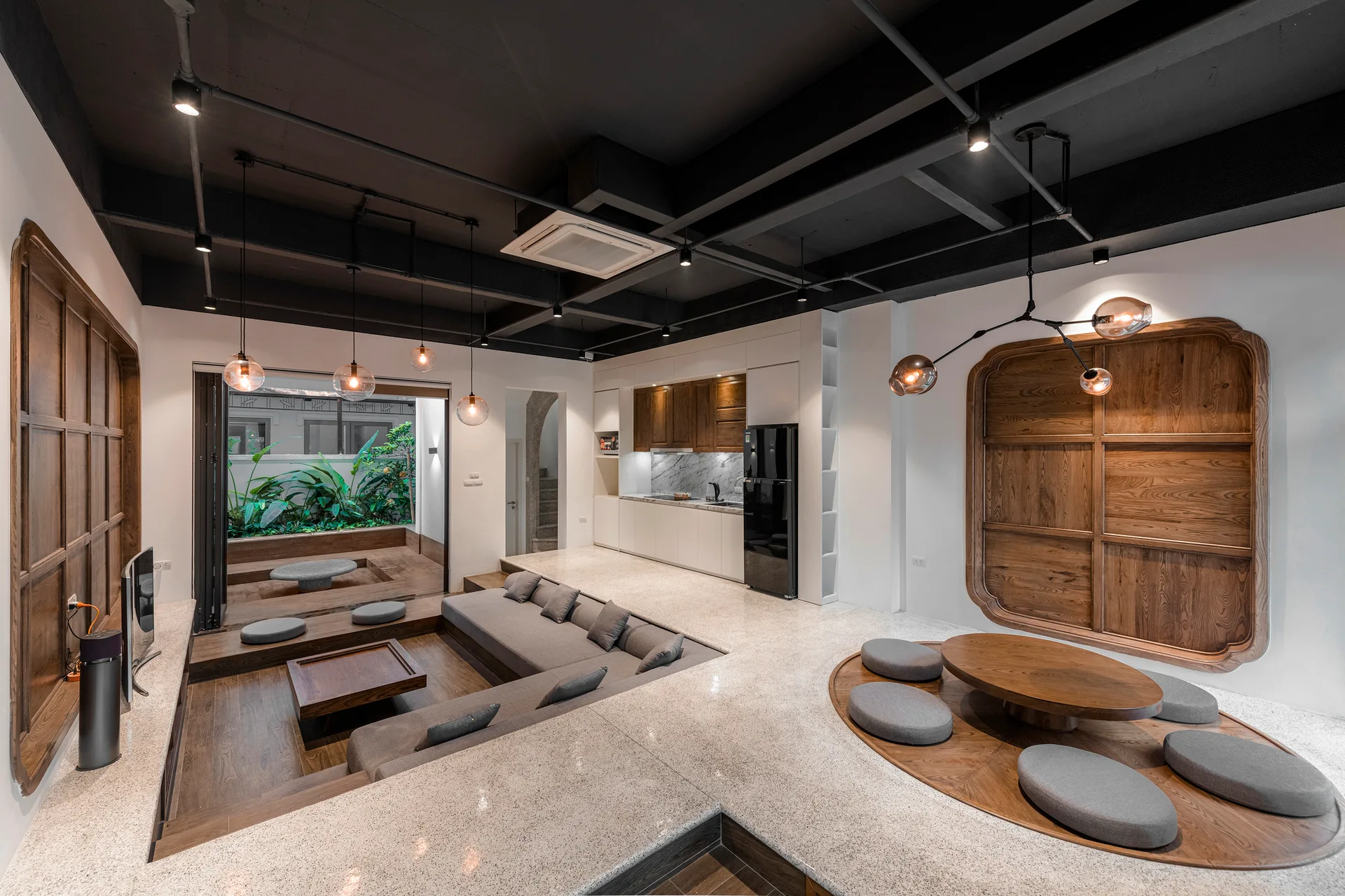 A modern open-concept living room features a sunken grey sectional sofa, low wooden dining table with floor cushions, and a black industrial ceiling.
