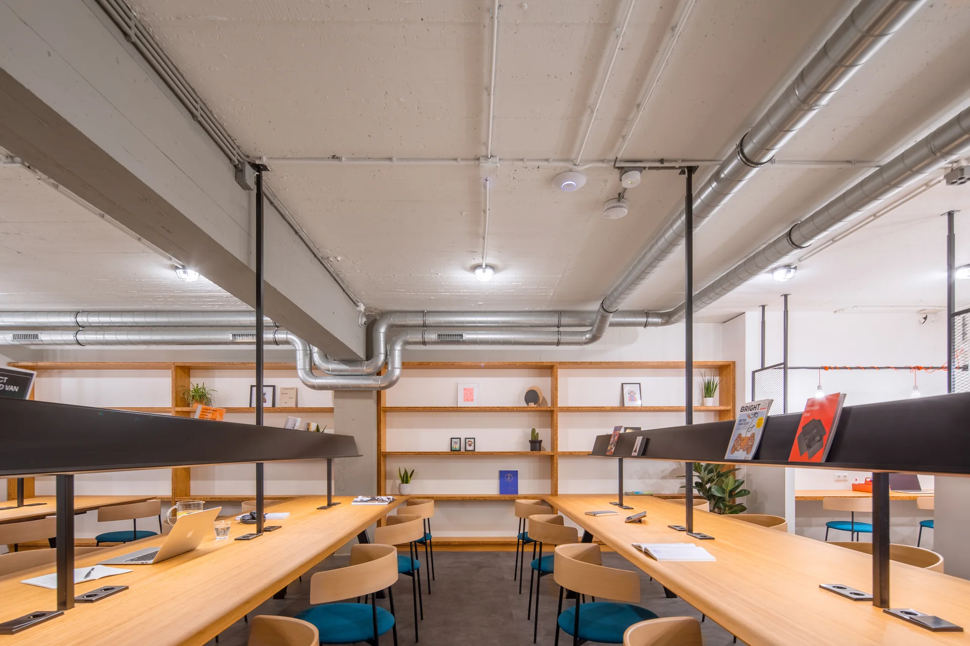 Long wooden communal tables with integrated black light fixtures and blue chairs sit under an exposed concrete ceiling with metal ductwork.