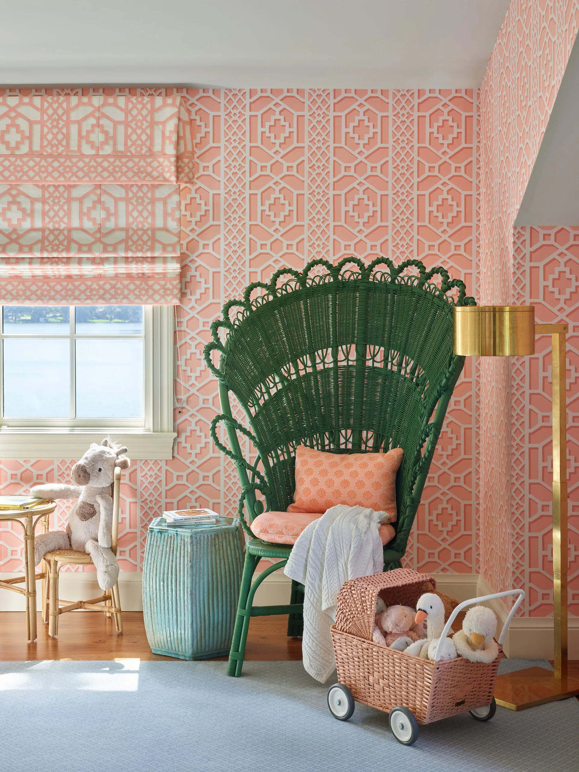 A green wicker peacock chair sits against peach and white geometric wallpaper next to a window with a matching roman shade in a children's room.