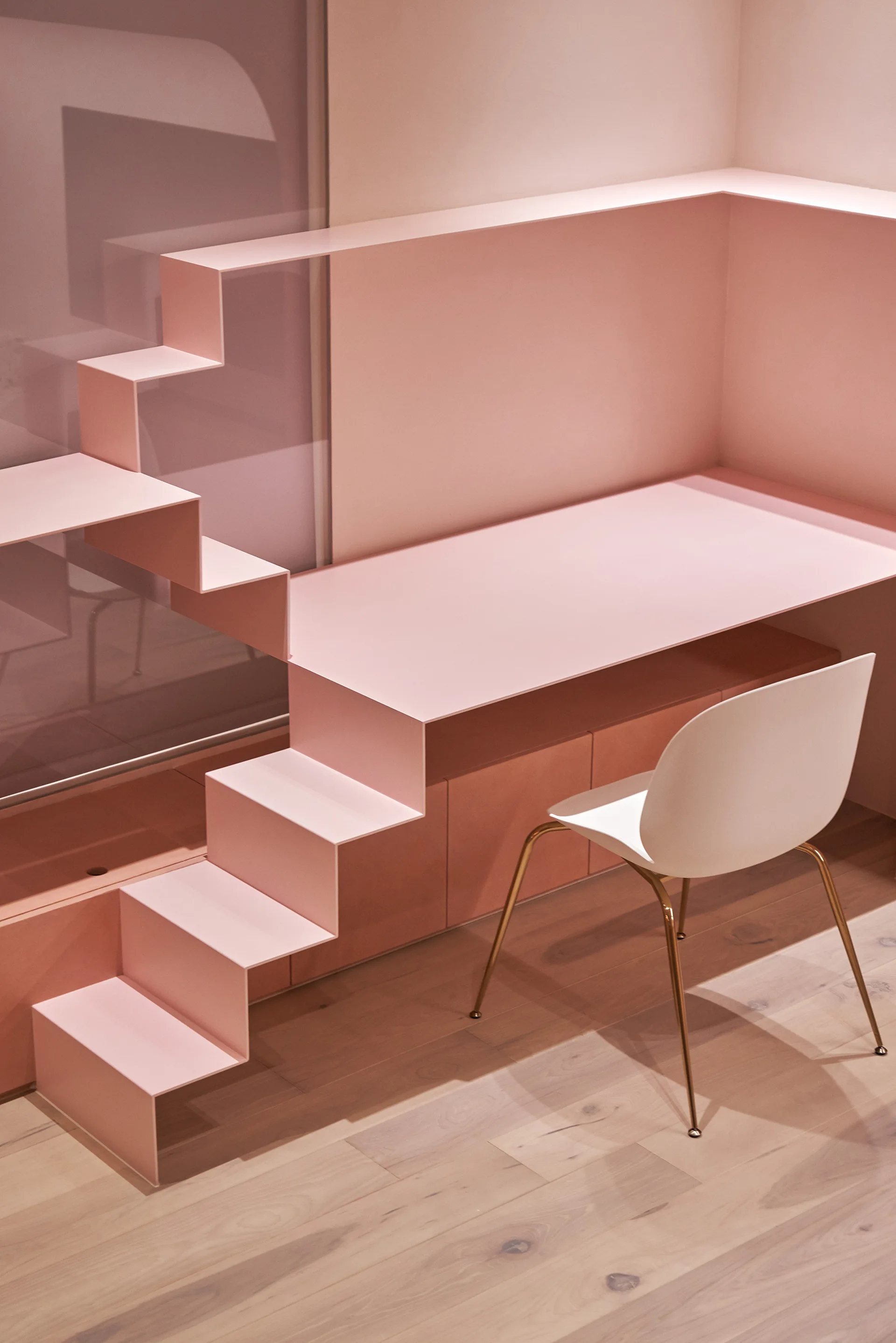 A modern home office workspace features a custom pink metal desk integrated with a geometric staircase next to a white chair with gold legs.