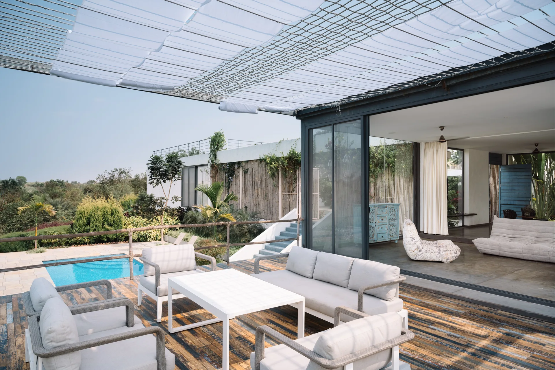 White outdoor lounge furniture sits on a weathered wood deck next to a swimming pool and large sliding glass doors leading inside.