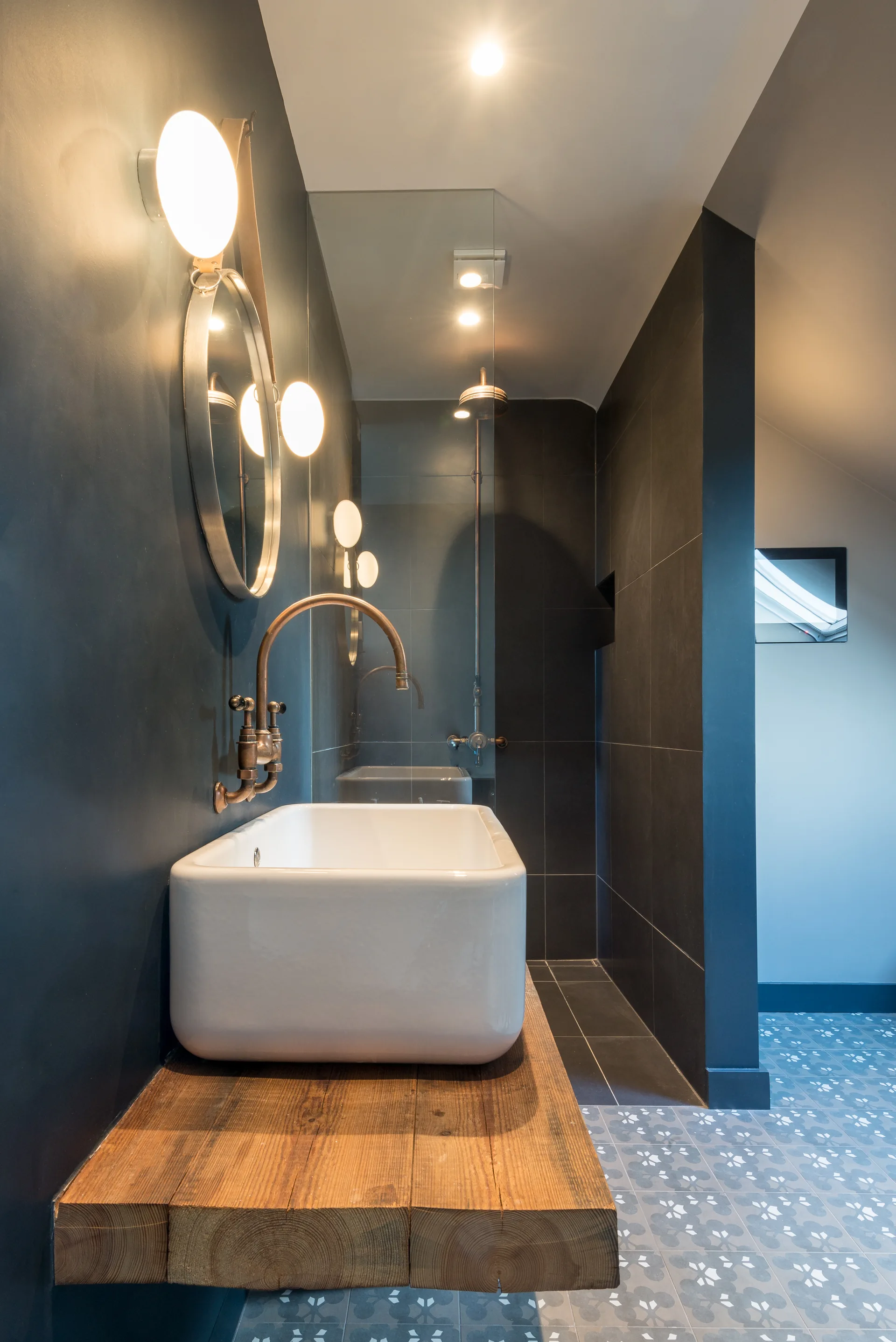 A white ceramic vessel sink sits on a thick timber slab vanity in a bathroom with dark charcoal walls, copper fixtures, and patterned floor tiles.