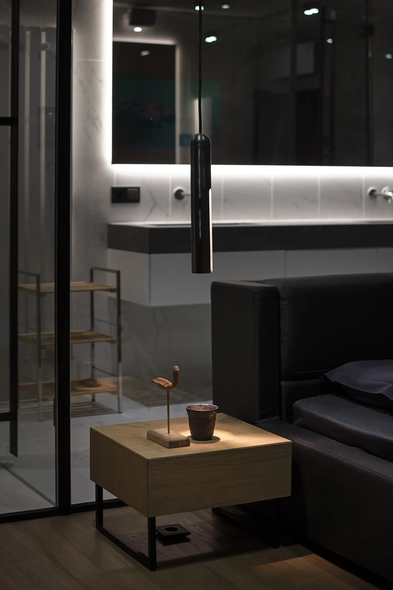 A wooden nightstand sits next to a black bed beneath a hanging black pendant light, with a glass partition showing a bathroom vanity in the background.
