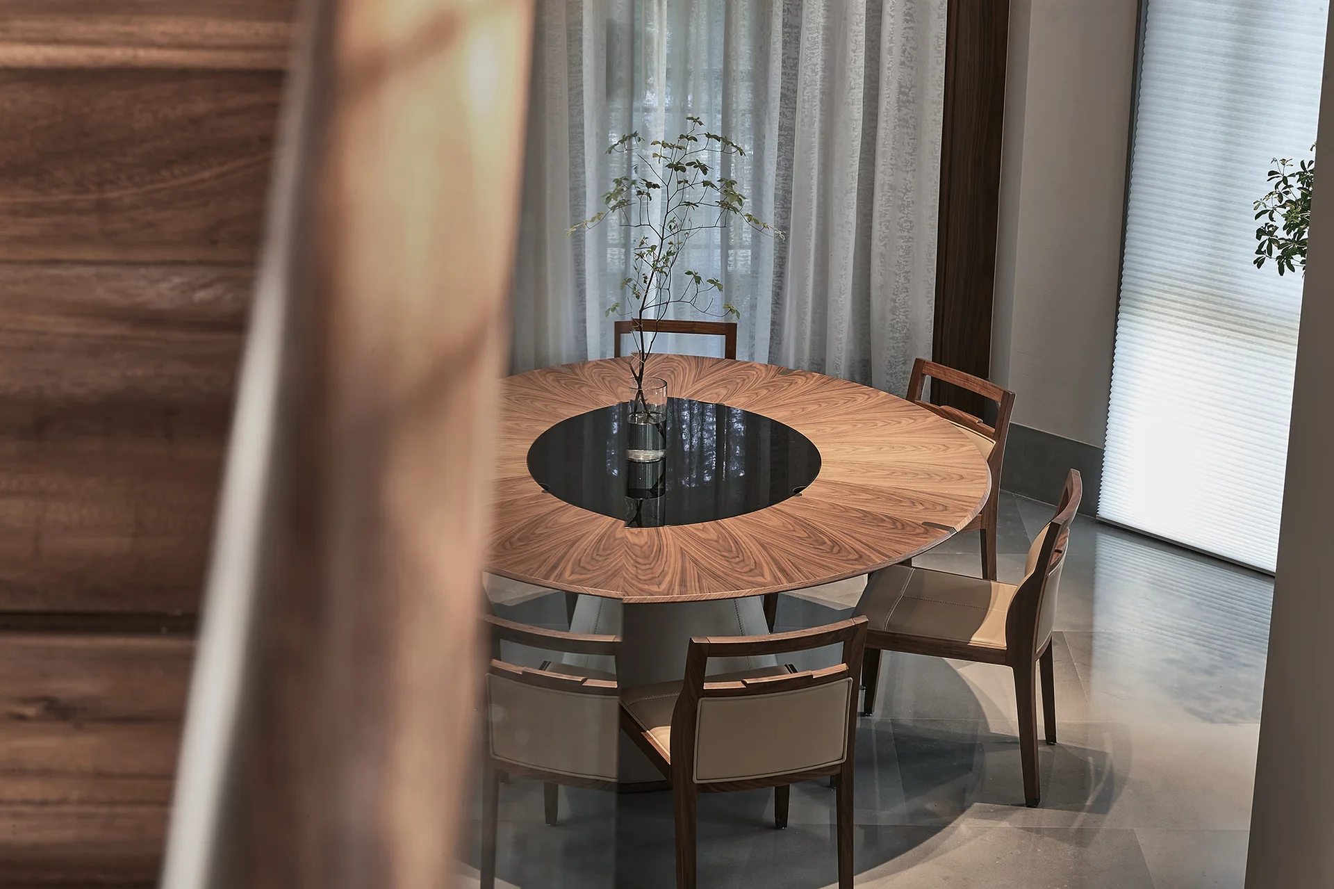 A circular wooden dining table with a black center inset and matching chairs stands in front of sheer white curtains.