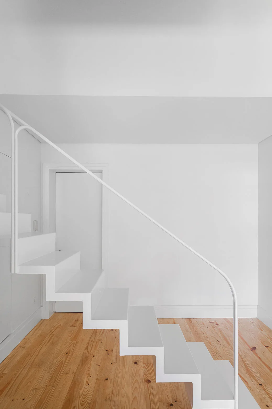A minimalist white staircase with a slim metal railing ascends over light pine wood plank flooring against clean white walls.