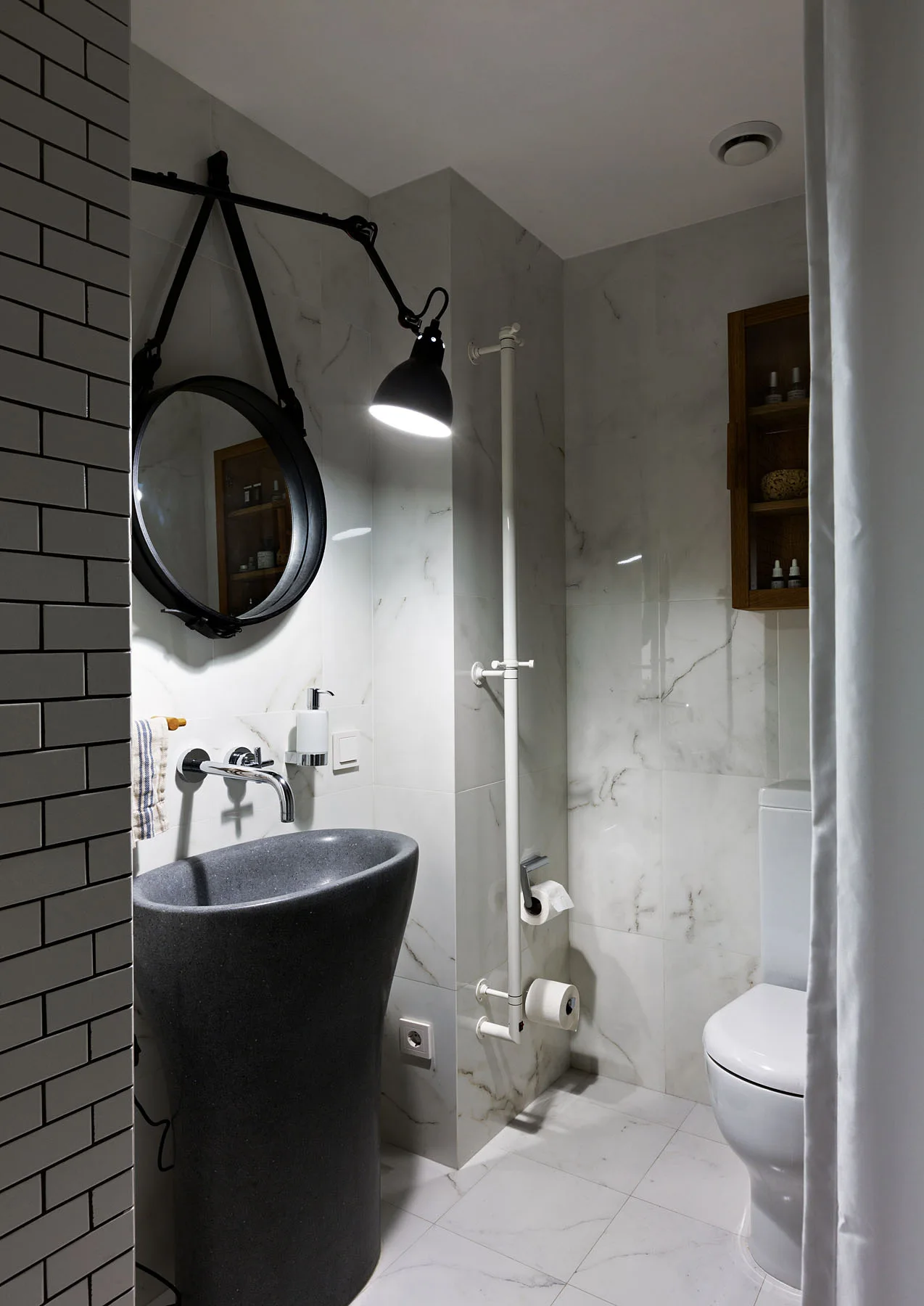 A modern bathroom features a dark grey pedestal sink, white marble wall tiles, a round black mirror, and a white toilet.