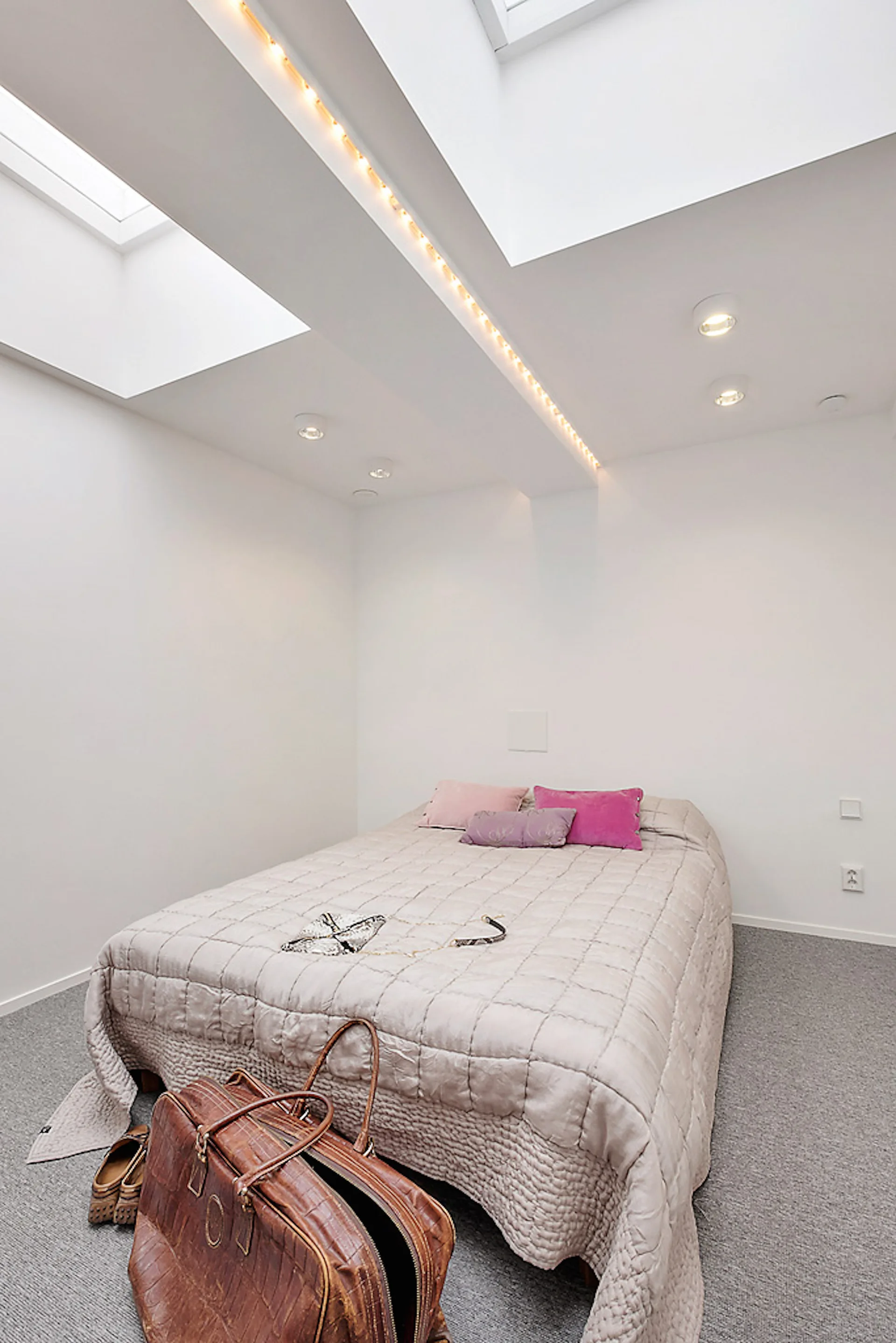 A minimalist attic bedroom features a bed with a quilted beige coverlet under a white ceiling with dual skylights, a center beam, and grey carpet.