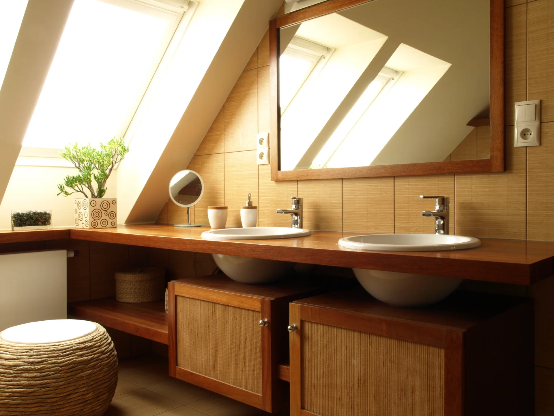 A bathroom vanity area featuring double vessel sinks, a large wood-framed mirror, and wood cabinets under a sloped ceiling with a bright skylight.