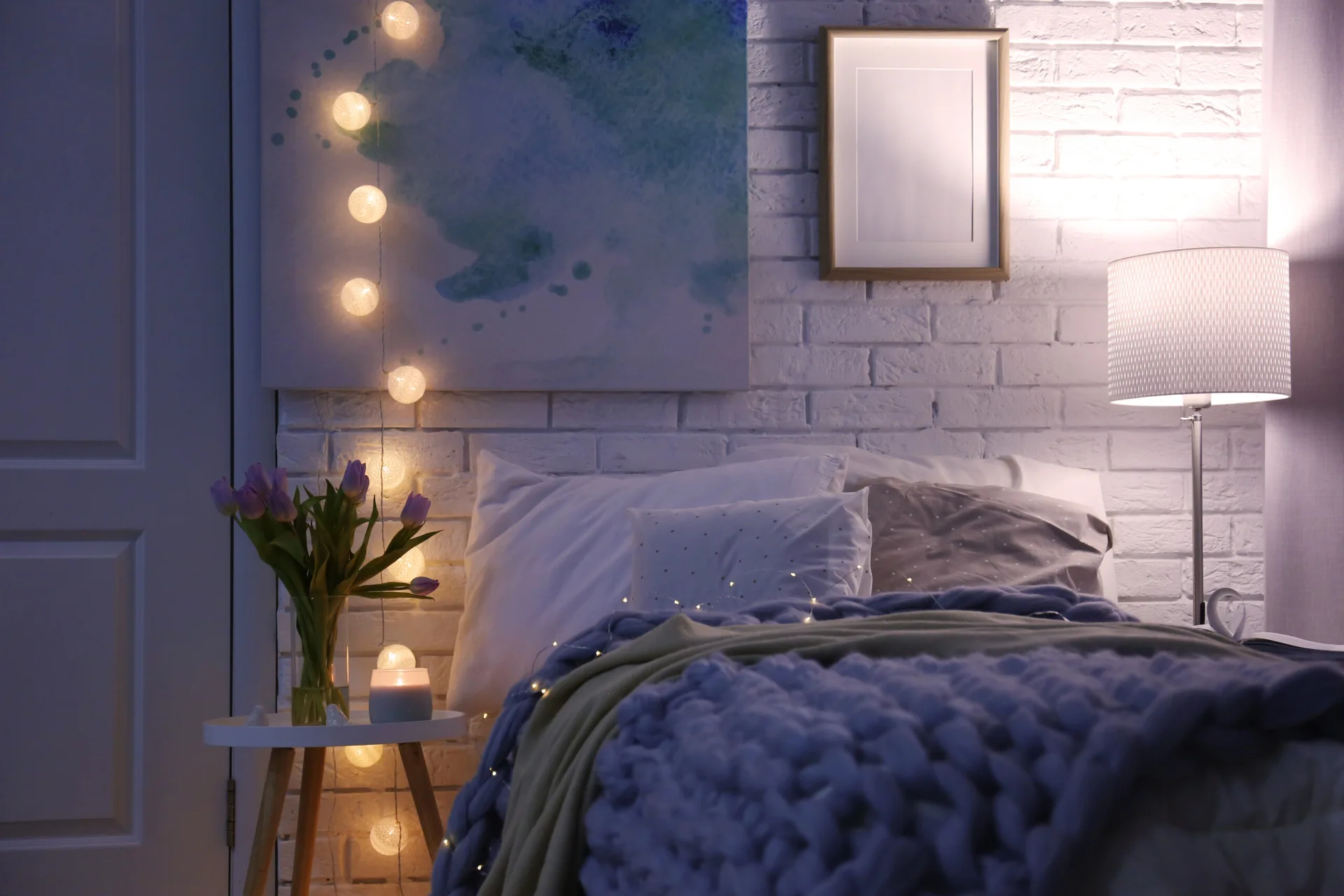 A bedroom features a white brick wall decorated with globe string lights, an abstract painting, and a bed with a chunky blue knit blanket.