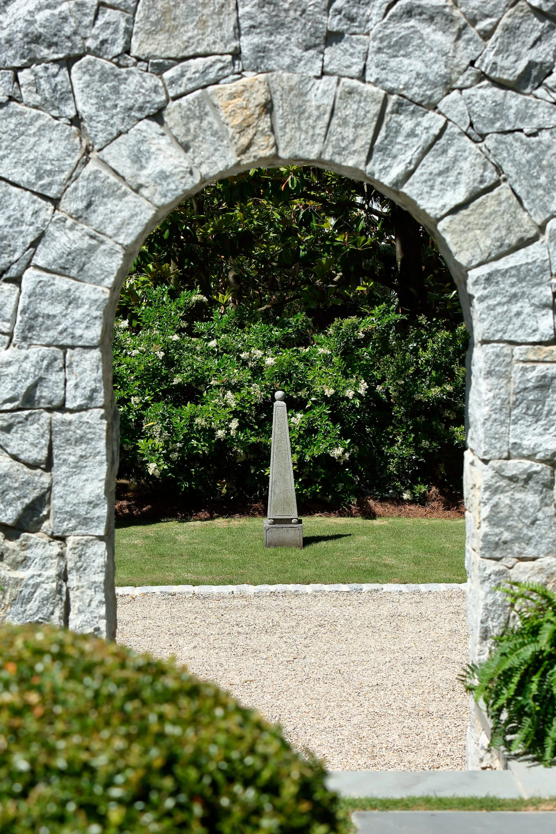 A rough grey stone archway frames a view of a manicured green lawn with a central stone obelisk sculpture and dense green shrubbery.