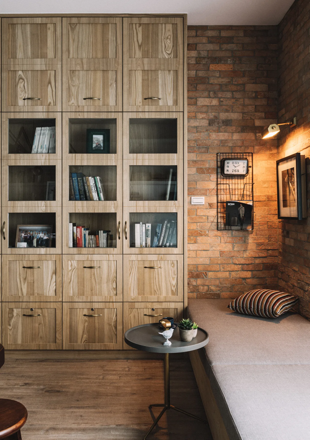 A reading nook featuring a floor-to-ceiling light wood storage cabinet with glass doors next to an upholstered bench against an exposed brick wall.