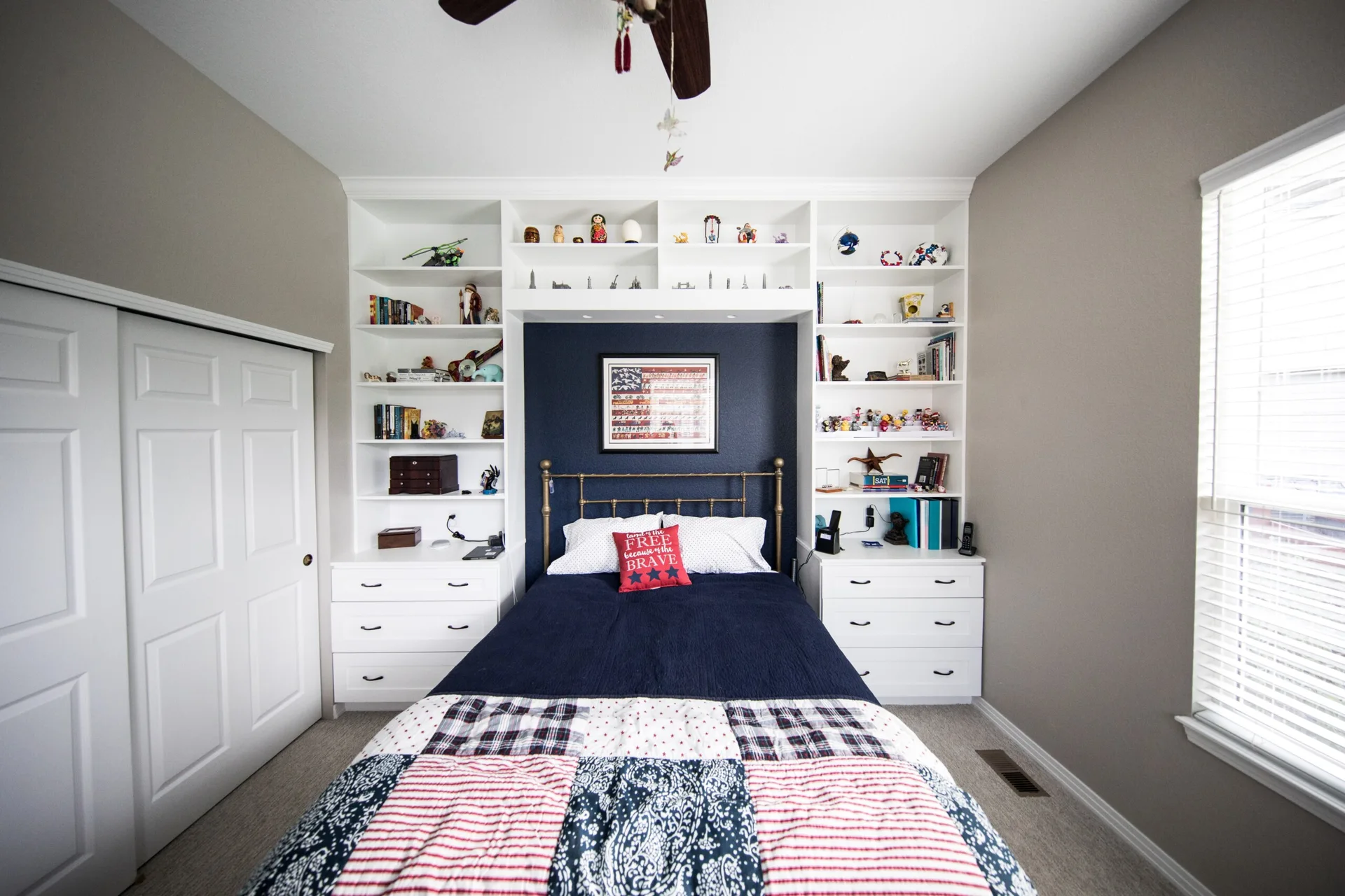 A bed with a brass frame and patchwork quilt is centered between symmetrical white built-in shelving units against a navy blue accent wall.