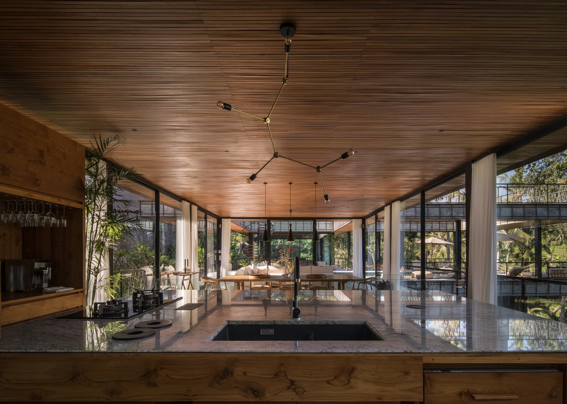 A modern kitchen featuring a polished stone island countertop under a slatted wood ceiling, with glass walls looking out to a green landscape.