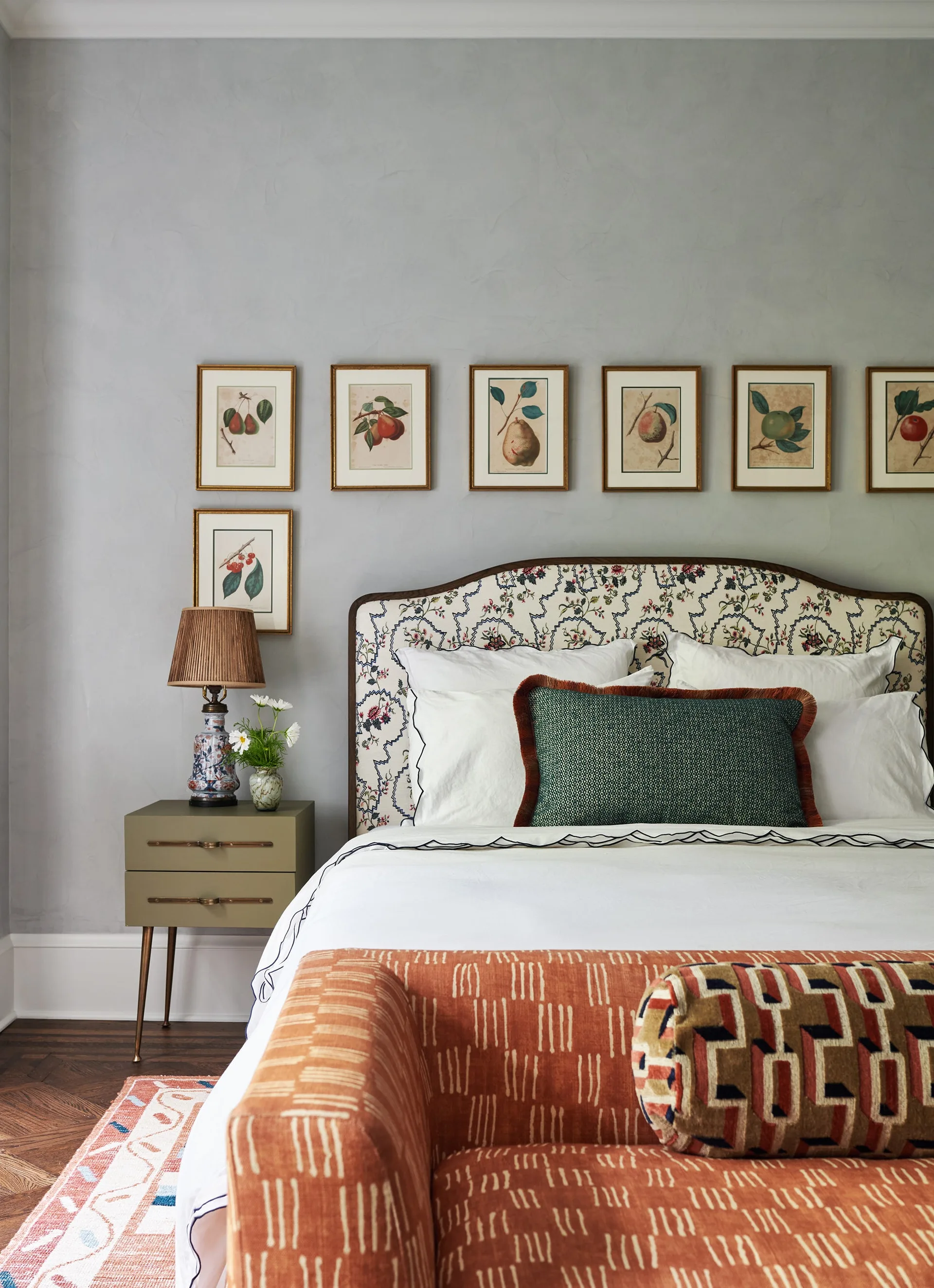 A bedroom features an upholstered floral headboard, a row of framed botanical prints, and an olive green nightstand against a textured grey wall.