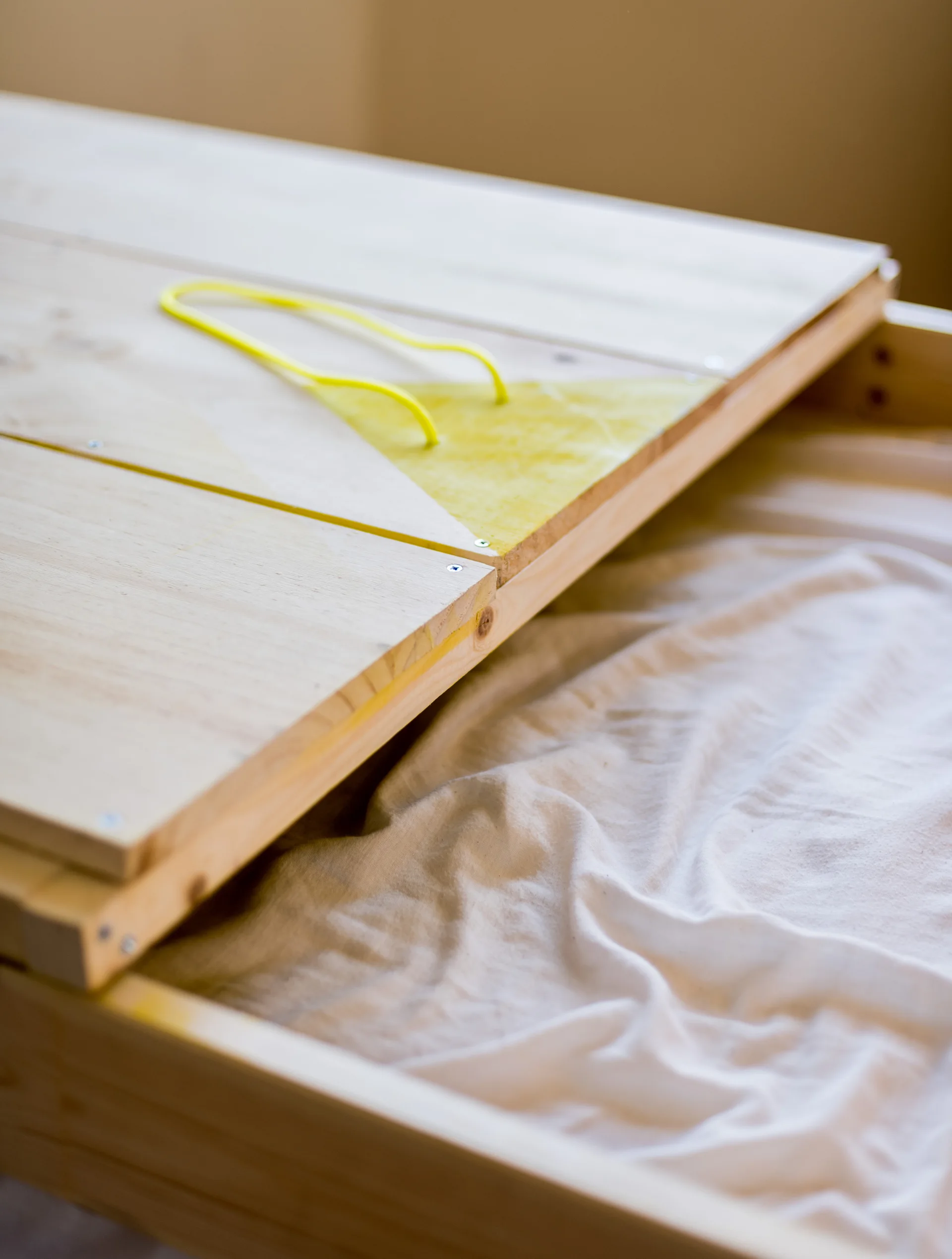 A close-up of an unfinished light wood storage drawer with a yellow cord handle on the lid and white fabric inside.