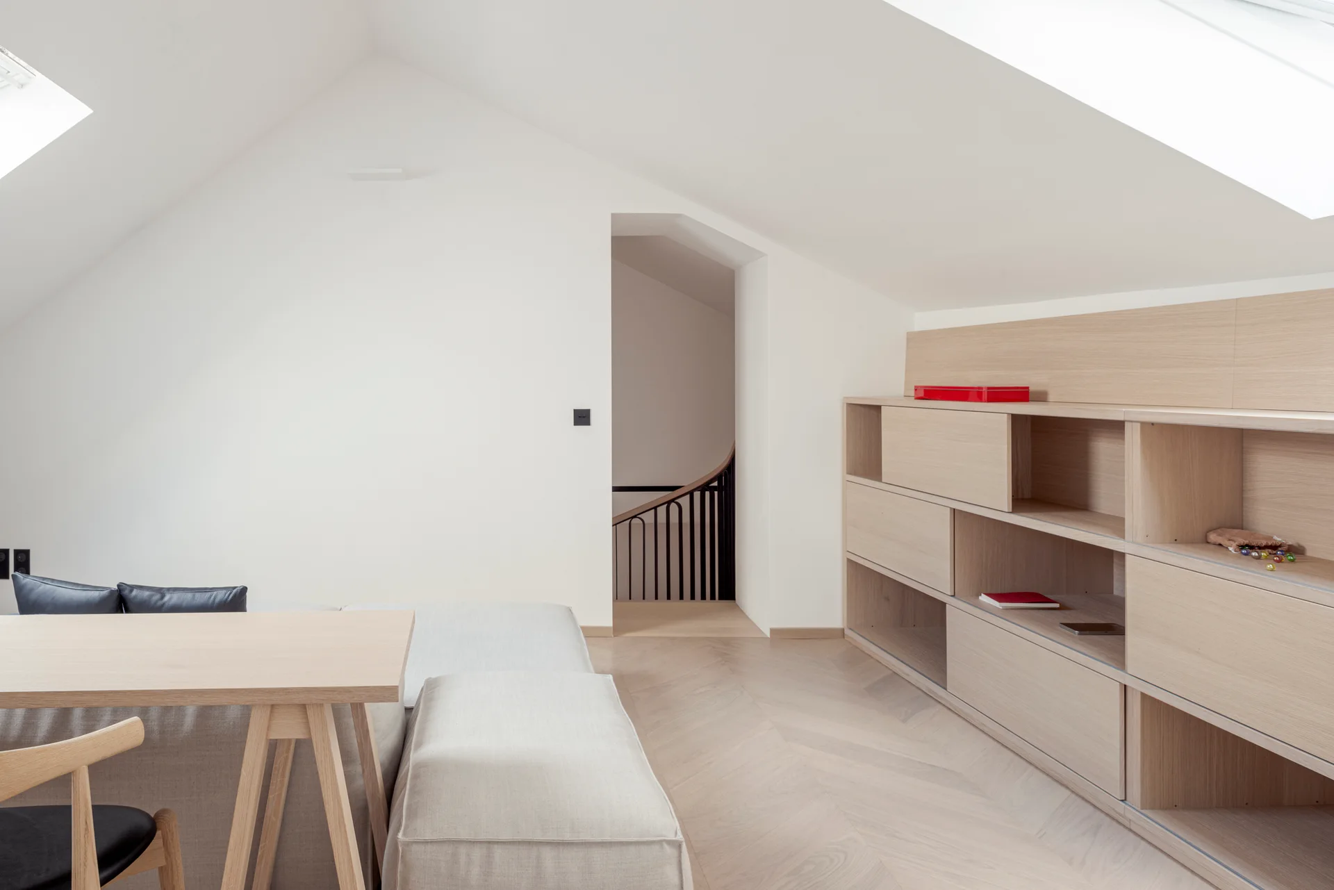 A bright attic room featuring custom light wood built-in shelving, chevron patterned flooring, a grey sofa, and skylights set in a sloped ceiling.