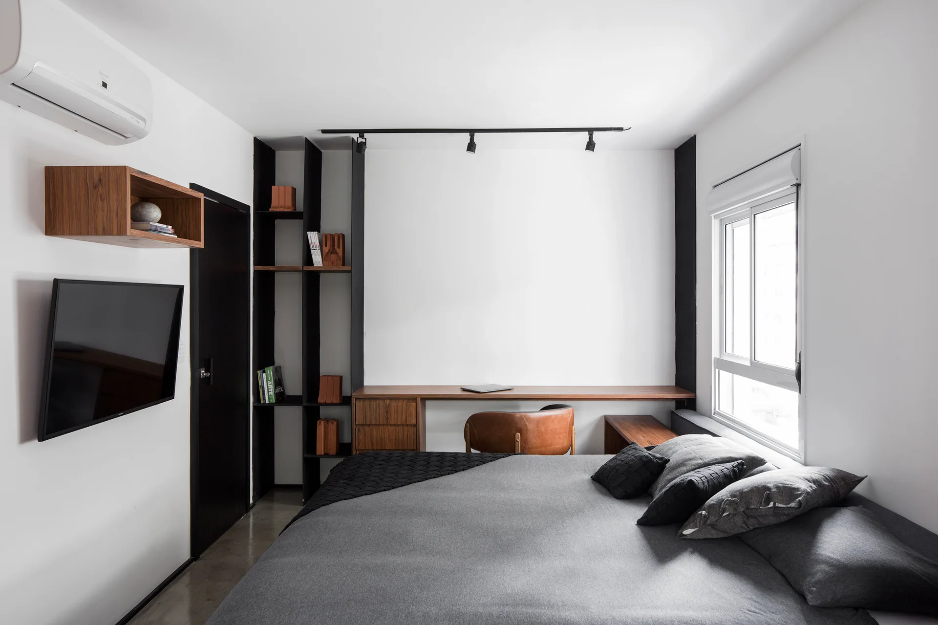 A minimalist bedroom features a grey-covered bed next to a built-in wooden desk, a tall black metal bookshelf, and ceiling track lighting.